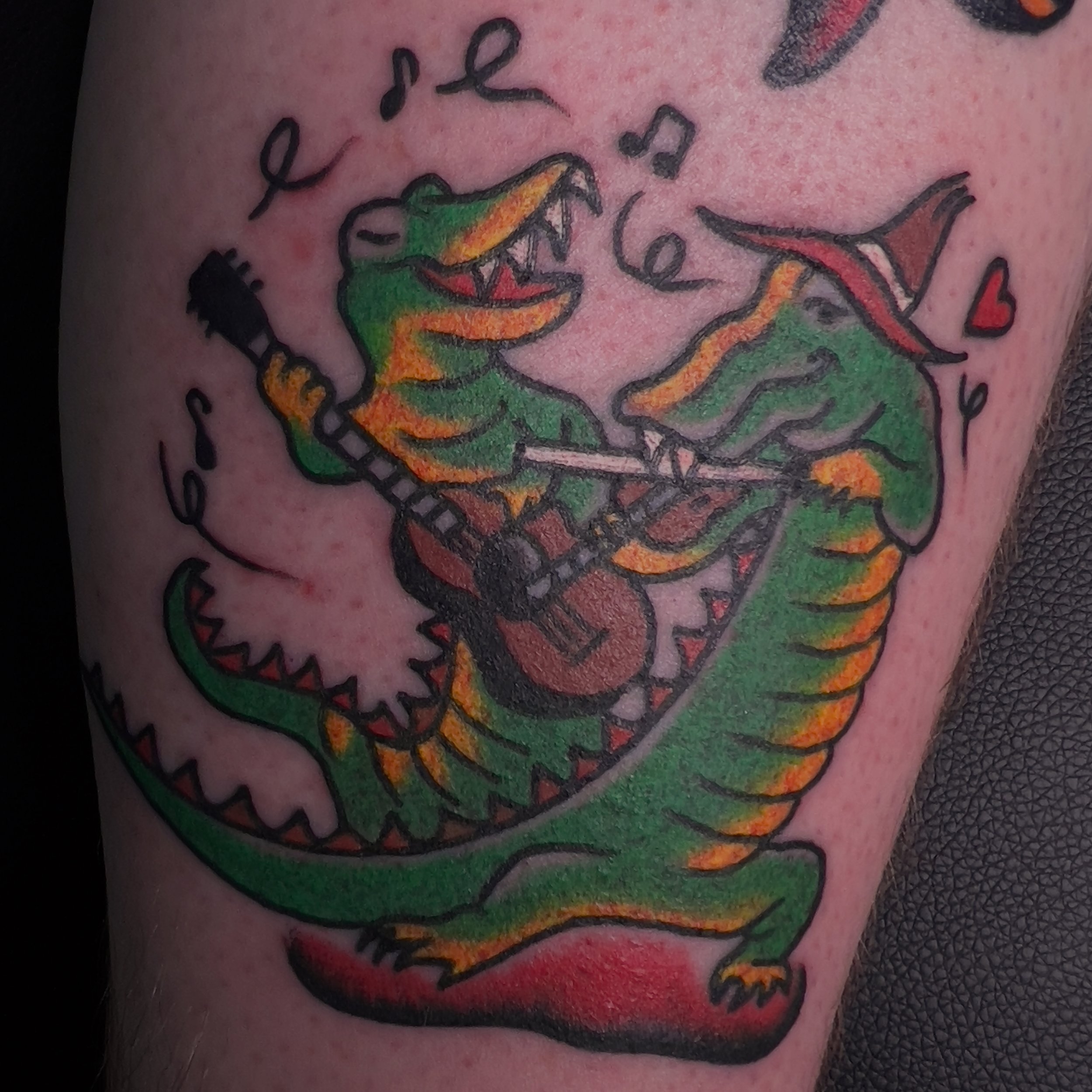 Tattoo of two cartoon crocodiles playing electric guitars, one wearing a red wizard hat, with musical notes and small hearts around them.