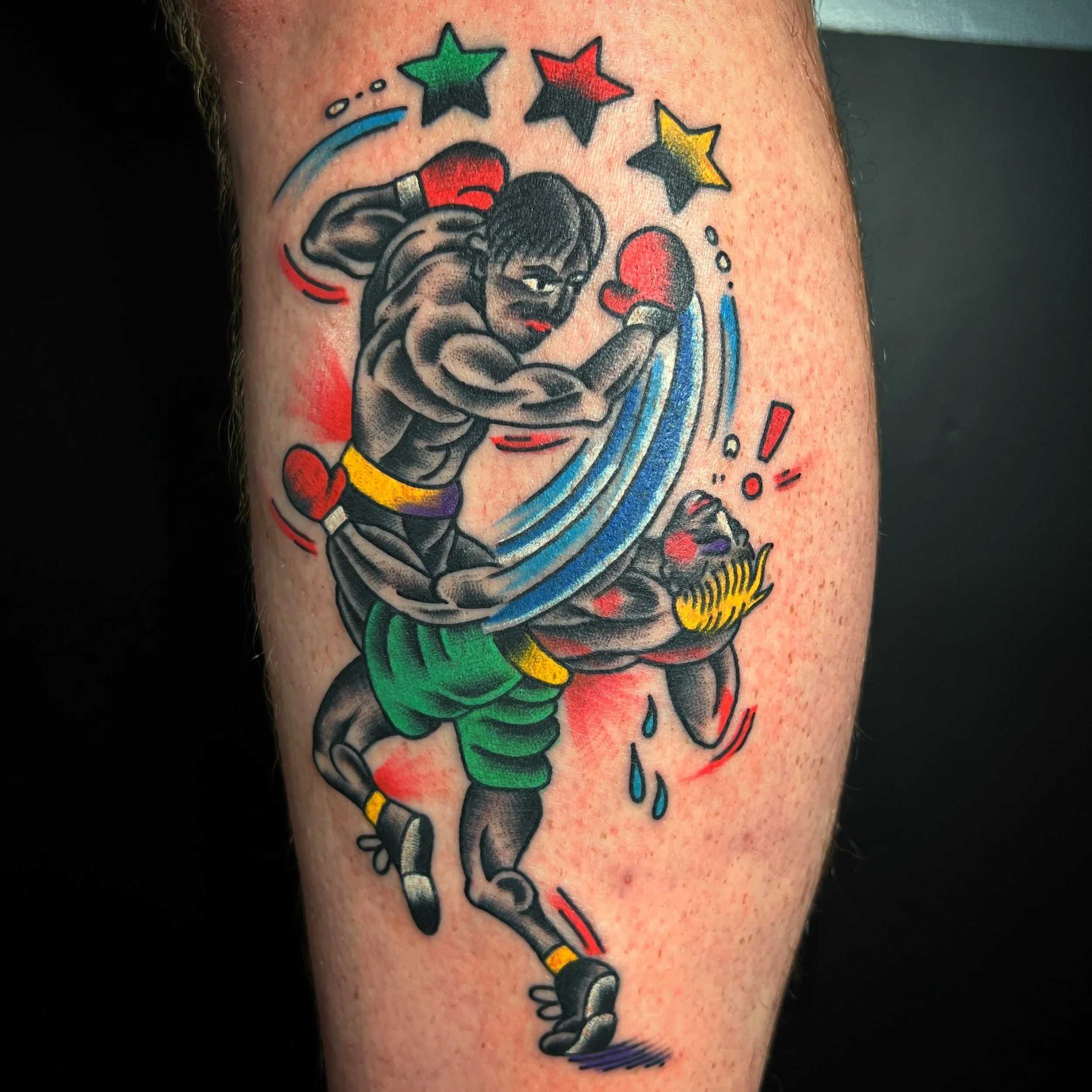 A colorful tattoo of two muscular boxers in a match, with one boxer punching the other. The tattoo features stars, hearts, and action lines, with red, green, yellow, blue, and black colors.