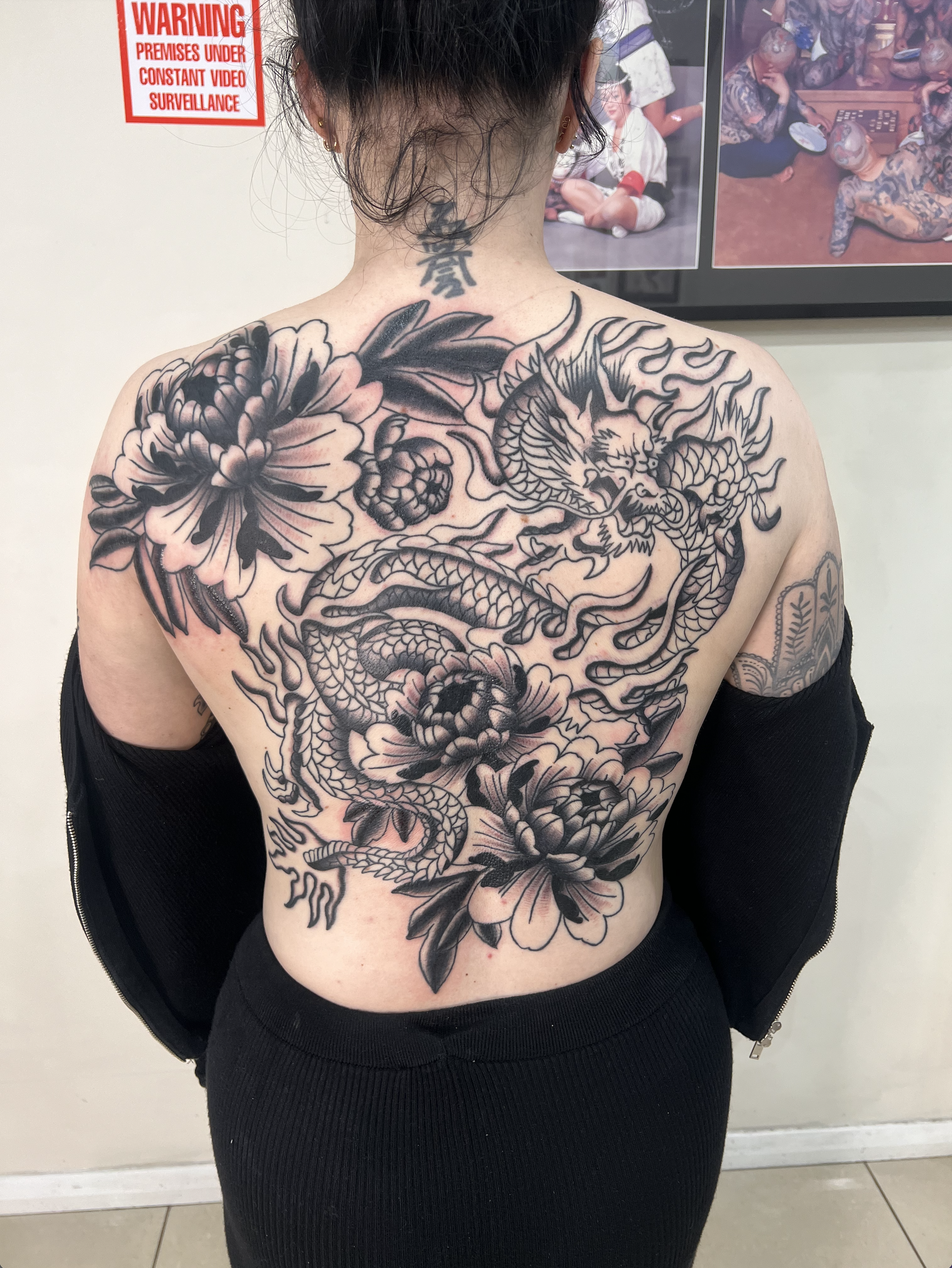 A woman with a large black and gray dragon and floral tattoo covering her back. She has a tattoo on her neck and is standing indoors near a wall with a red warning sign and photos.