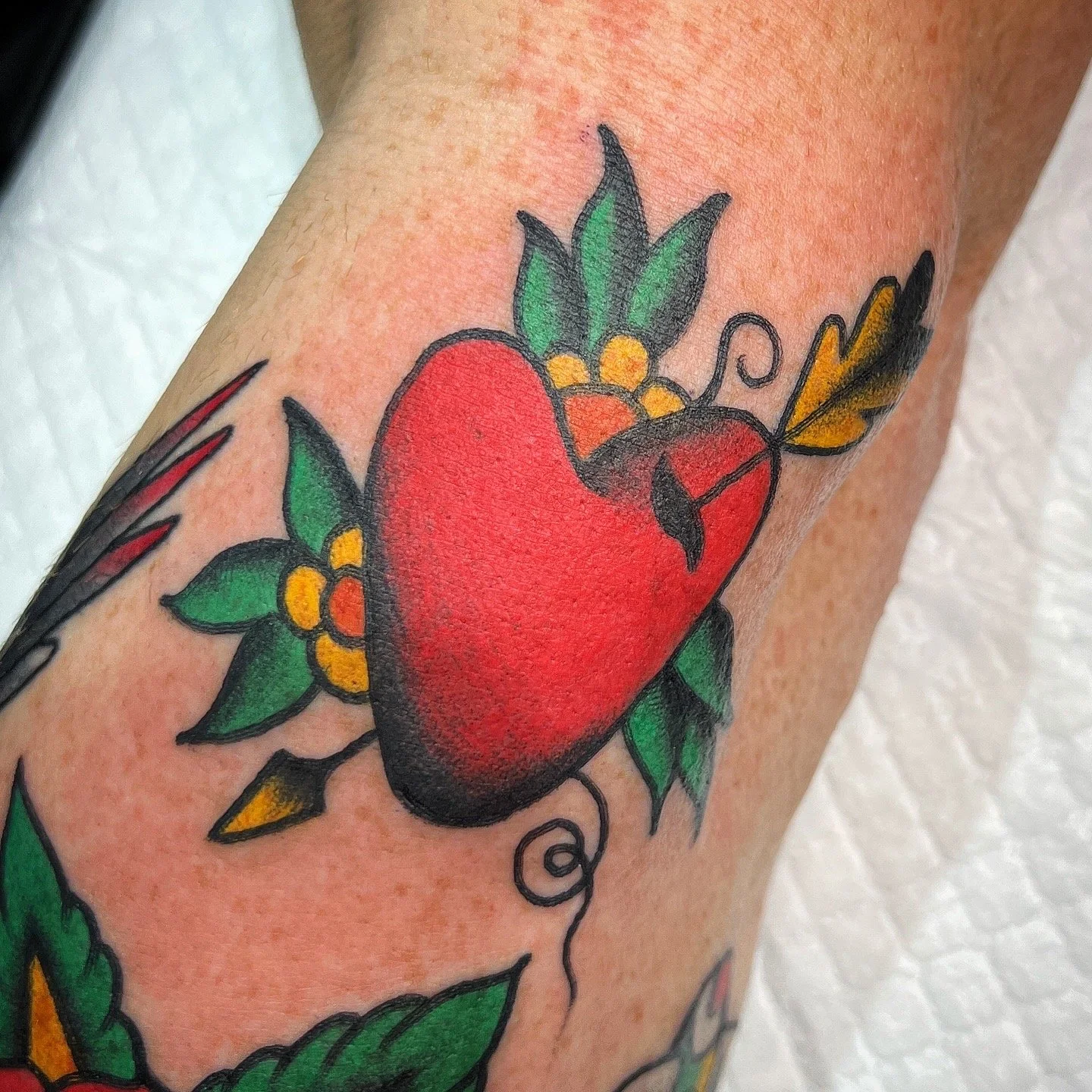 Tattoo of a red heart with green leaves and yellow-orange flowers around it.