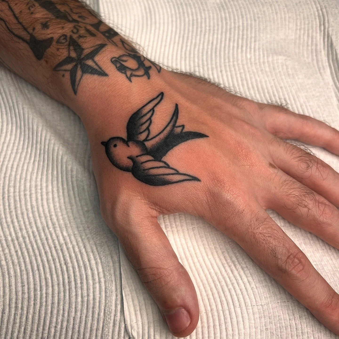 Black tattoo of a flying bird on the person's hand and wrist, with additional tattoos visible on the arm.