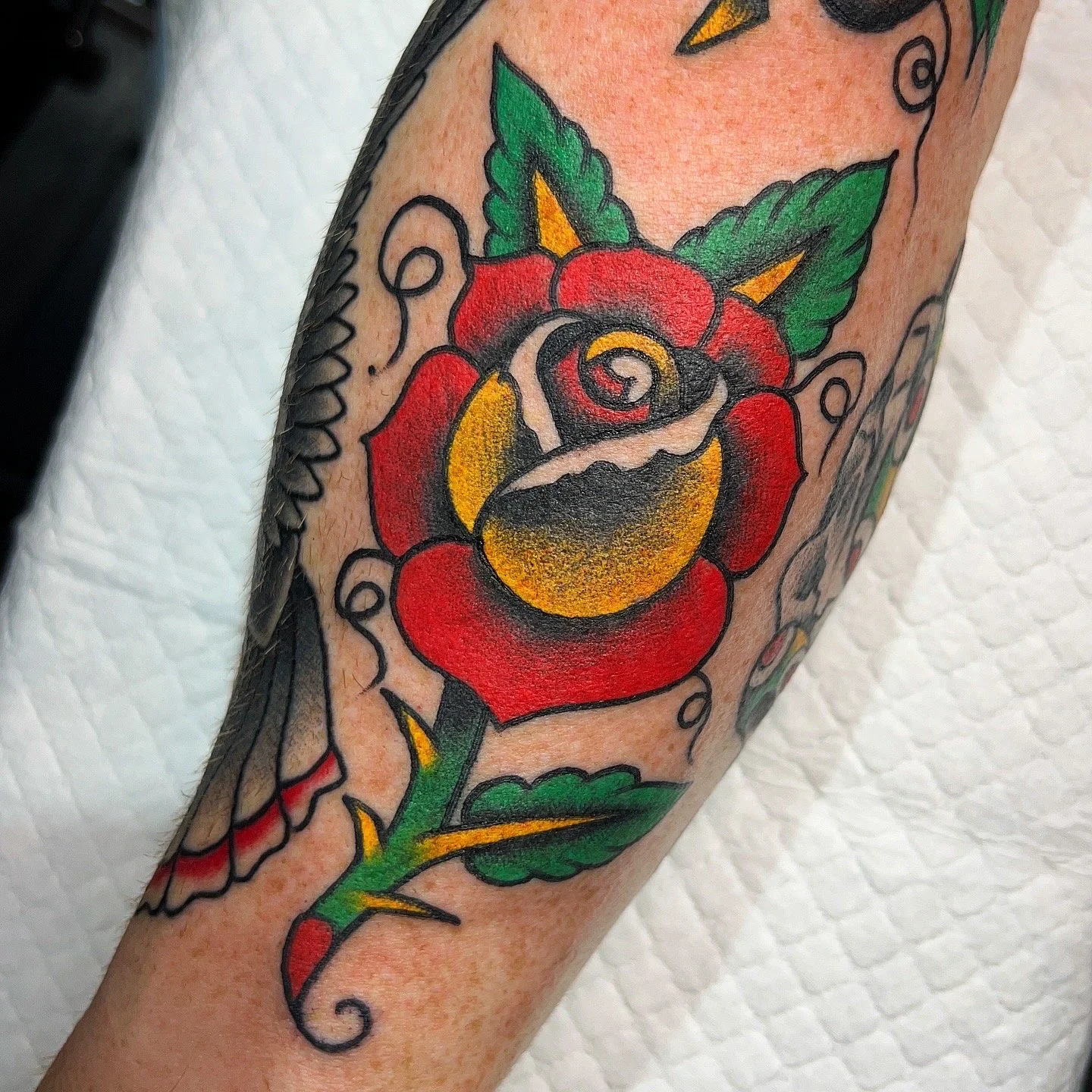Colorful tattoo of a traditional-style rose with green leaves and decorative black swirling lines.