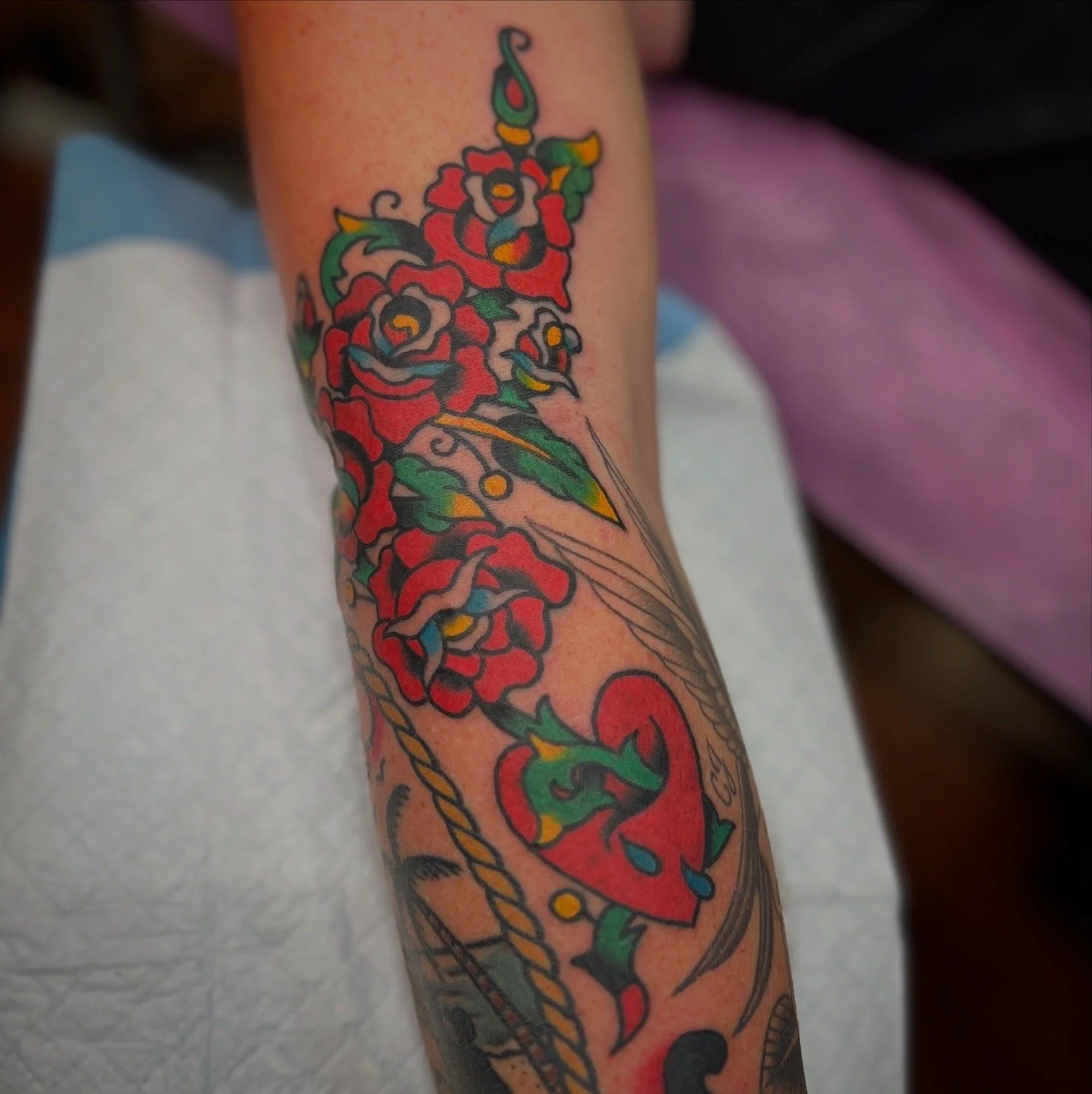 A tattoo of red roses, a green snake, a red heart, and a yellow and green dagger on a person's arm.