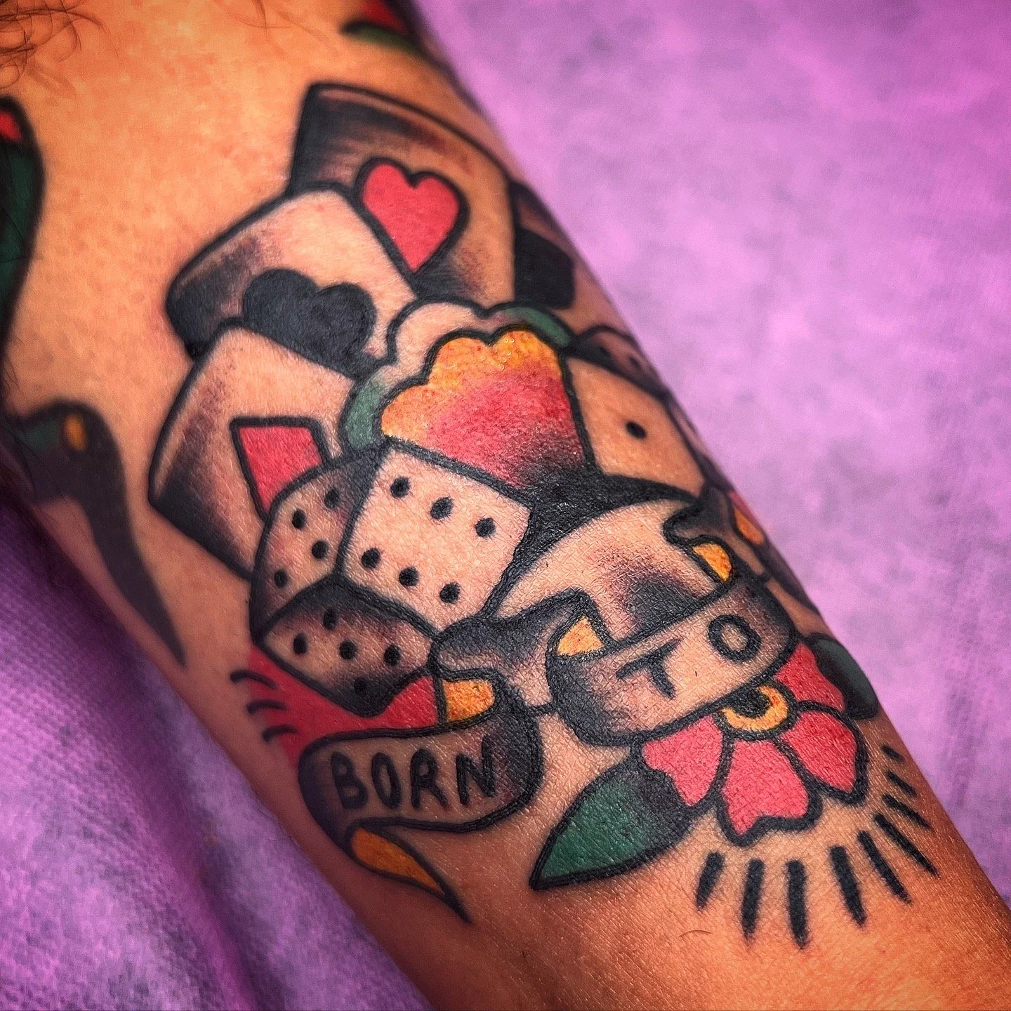 Tattoo of colored dice, hearts, and flowers with a banner that says 'BORN' on a person's arm.