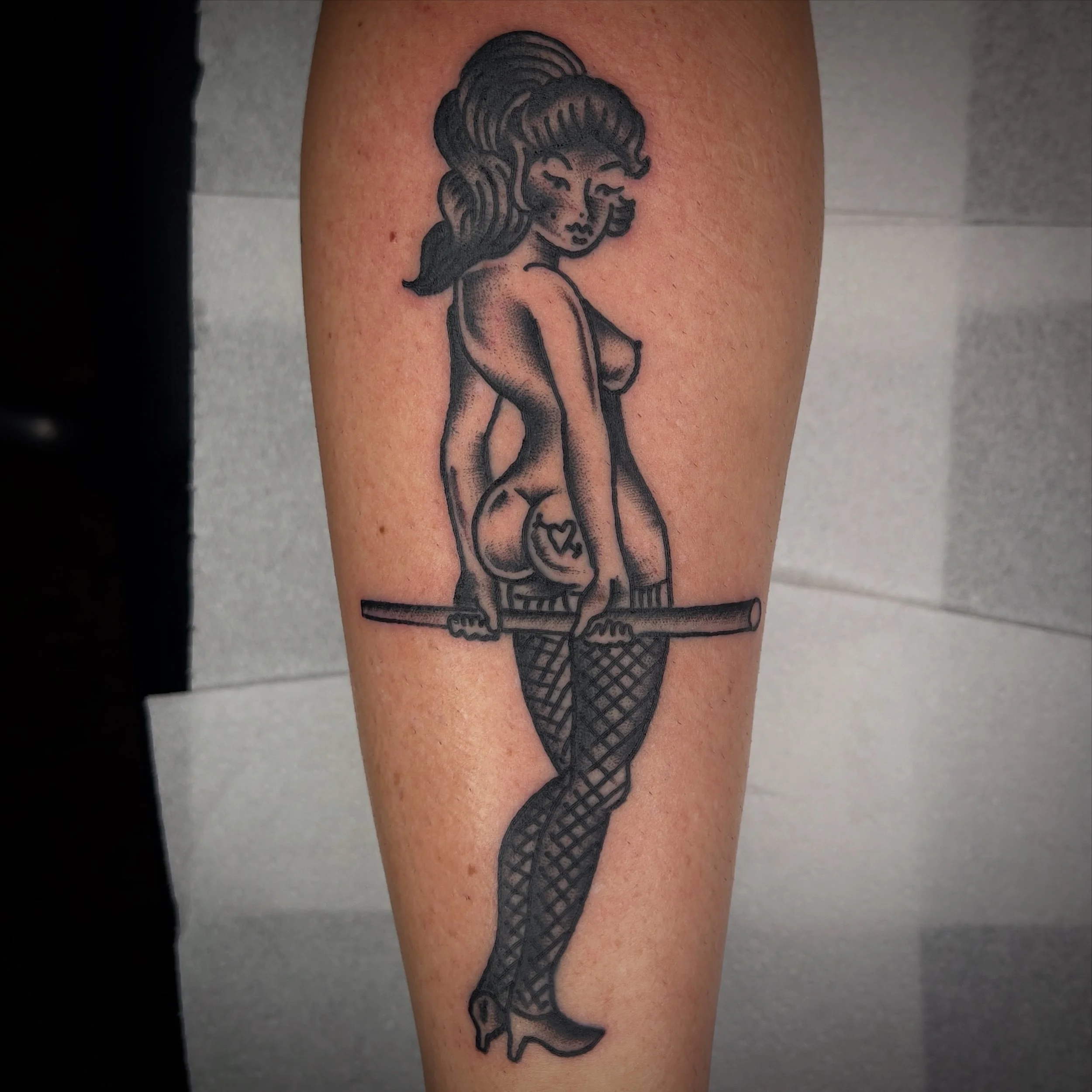 Tattoo of a caricature-style woman with curly hair, nude, sitting on a bar, wearing fishnet stockings and high heels.