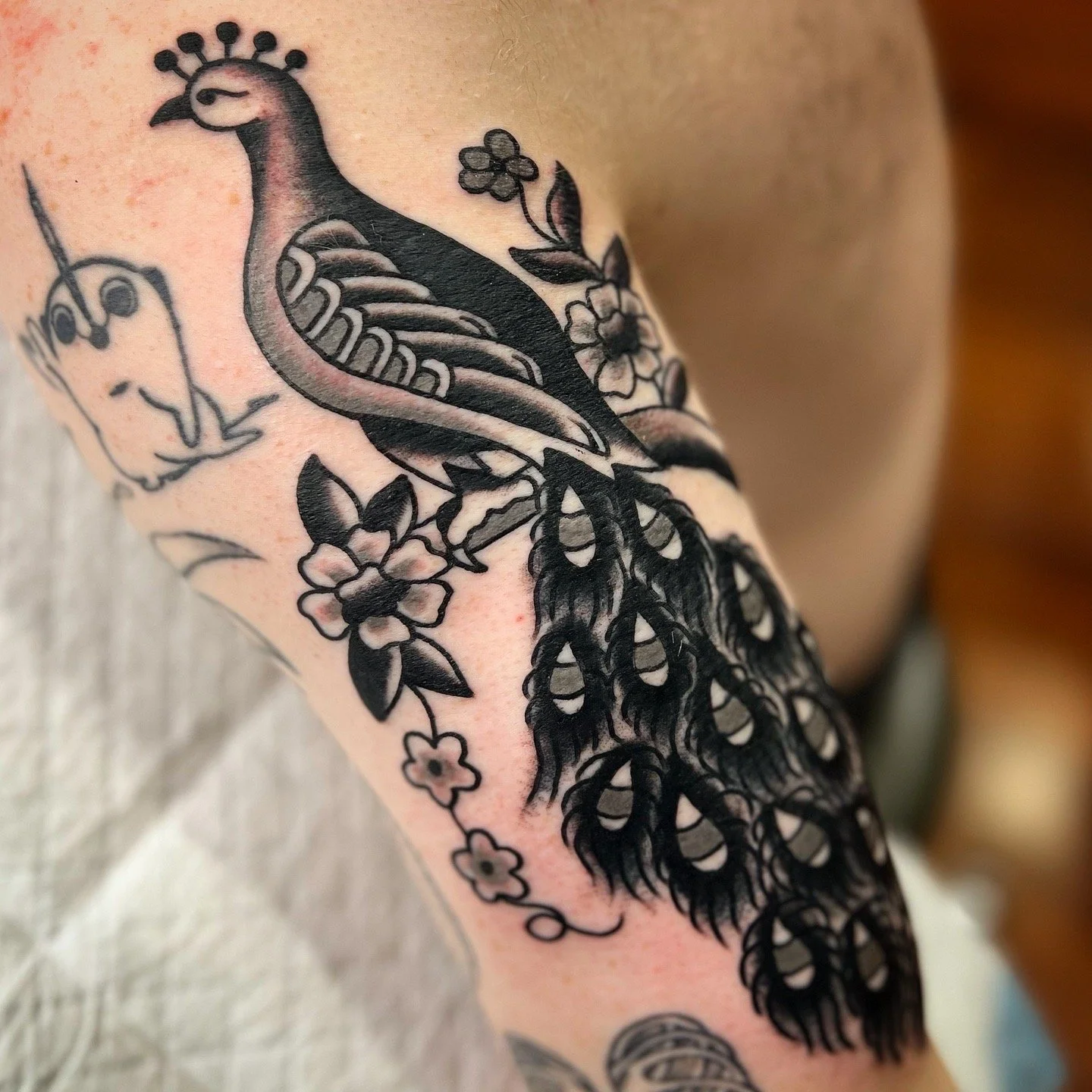 A black and gray tattoo of a peacock with floral details on a person's arm.