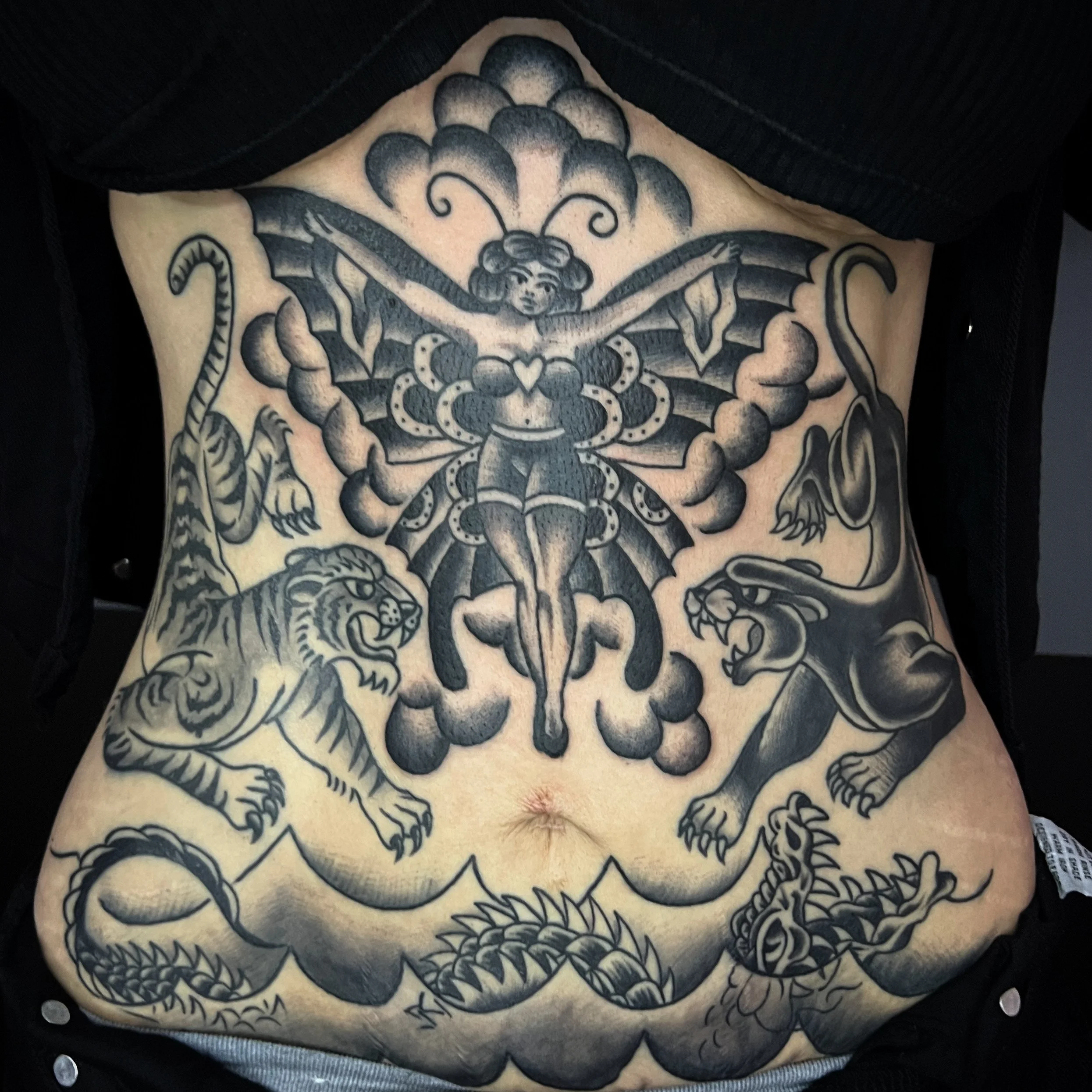 Black and gray tattoo on a person's stomach featuring a fairy with wings and a crown, flanked by a tiger on the left and a panther on the right, with a snake below.