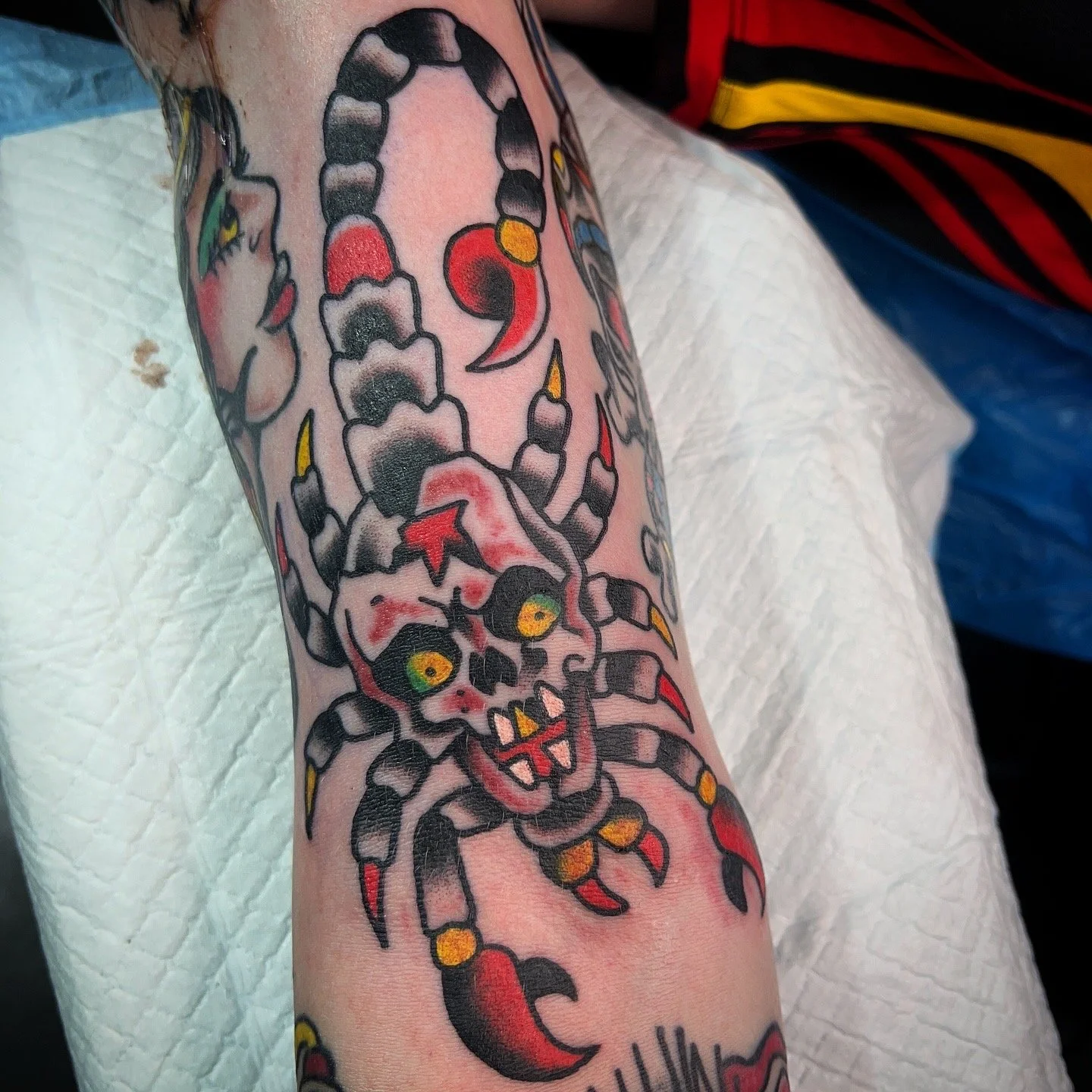 Tattoo of a skull with yellow eyes, jagged teeth, and red details, combined with a spider with red, black, and yellow accents, on a person's arm.