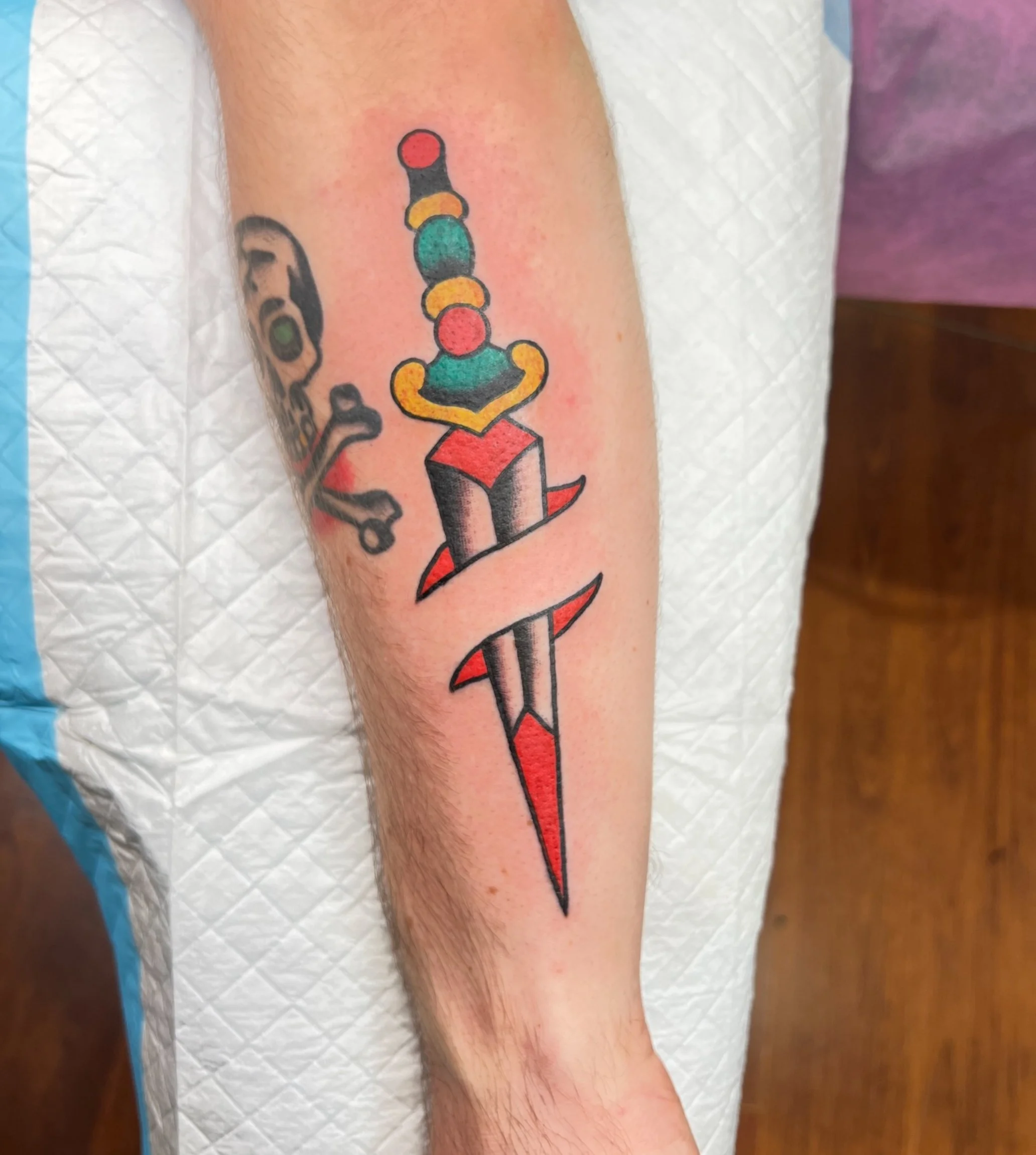 A tattoo of a colorful, stylized sword with a pointed red tip and a black and red handle on a person's arm.