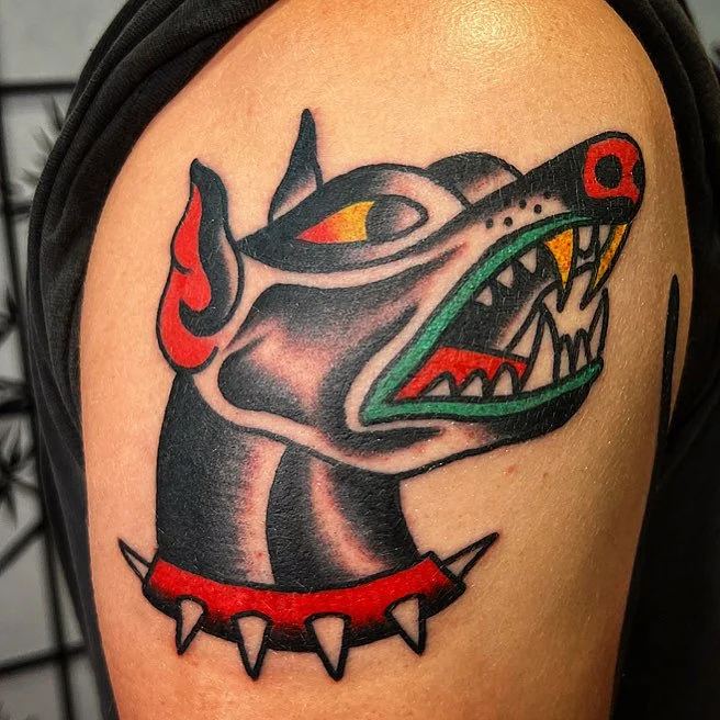 Tattoo of a stylized wolf head with a spiked collar, featuring bold black outlines, red, yellow, and green details on the face, and an aggressive expression with sharp teeth.