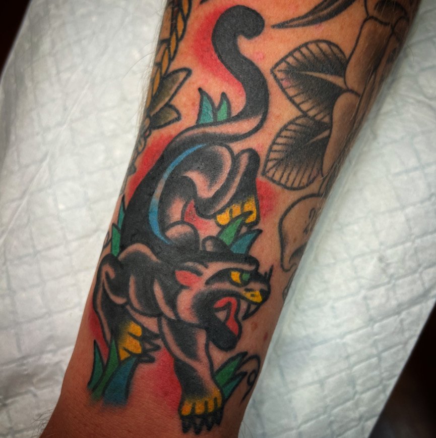 Tattoo of a fierce black panther with yellow eyes, surrounded by green grass, on a person's forearm.