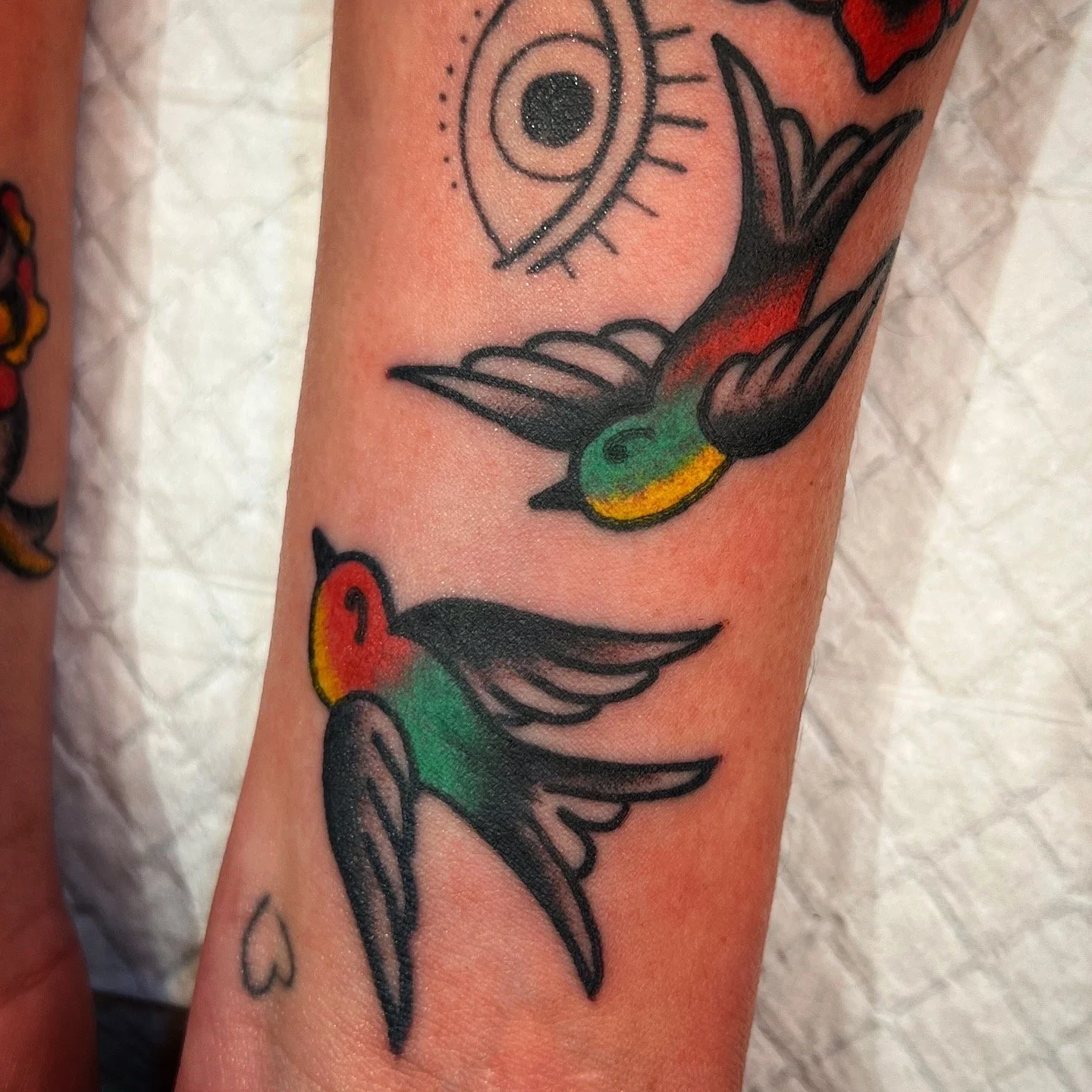 Tattoo of two colorful hummingbirds with green bodies and red heads flying near a sun with an eye design, all on person's arm.