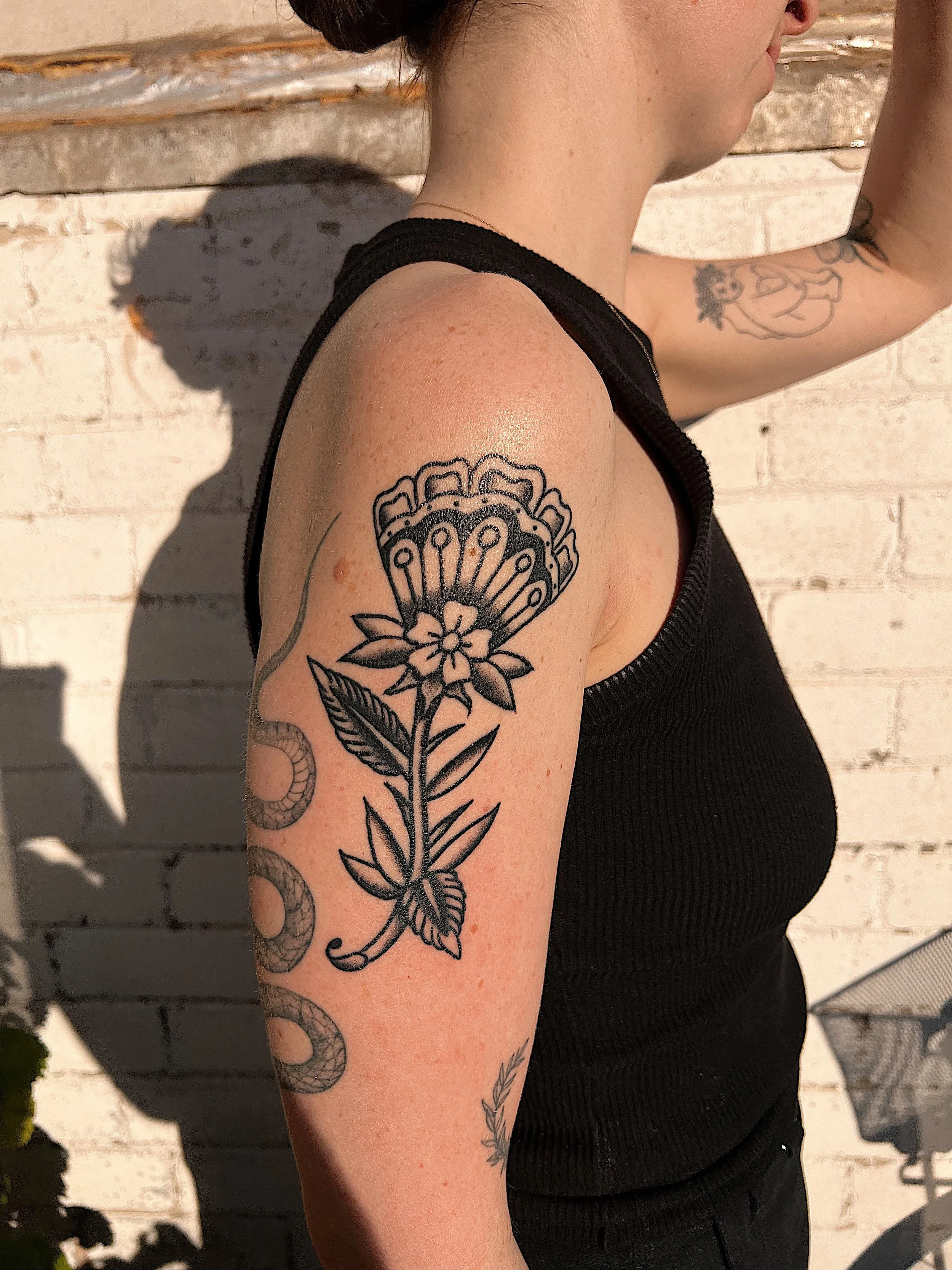 A woman with a tattoo of a flower on her upper arm, wearing a black sleeveless top, standing against a white brick wall.