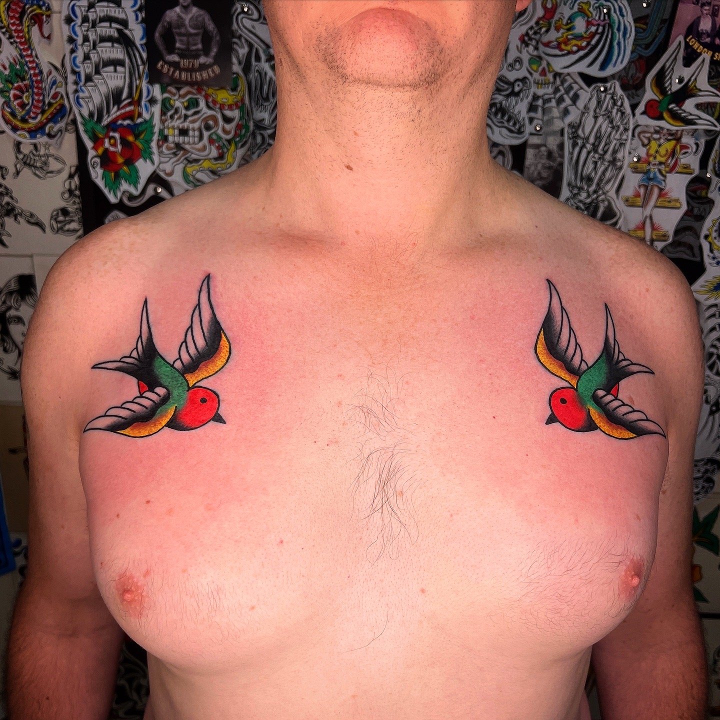 A person with two colorful swallow tattoos on their upper chest, near the shoulders, with a background of tattoo flash art.