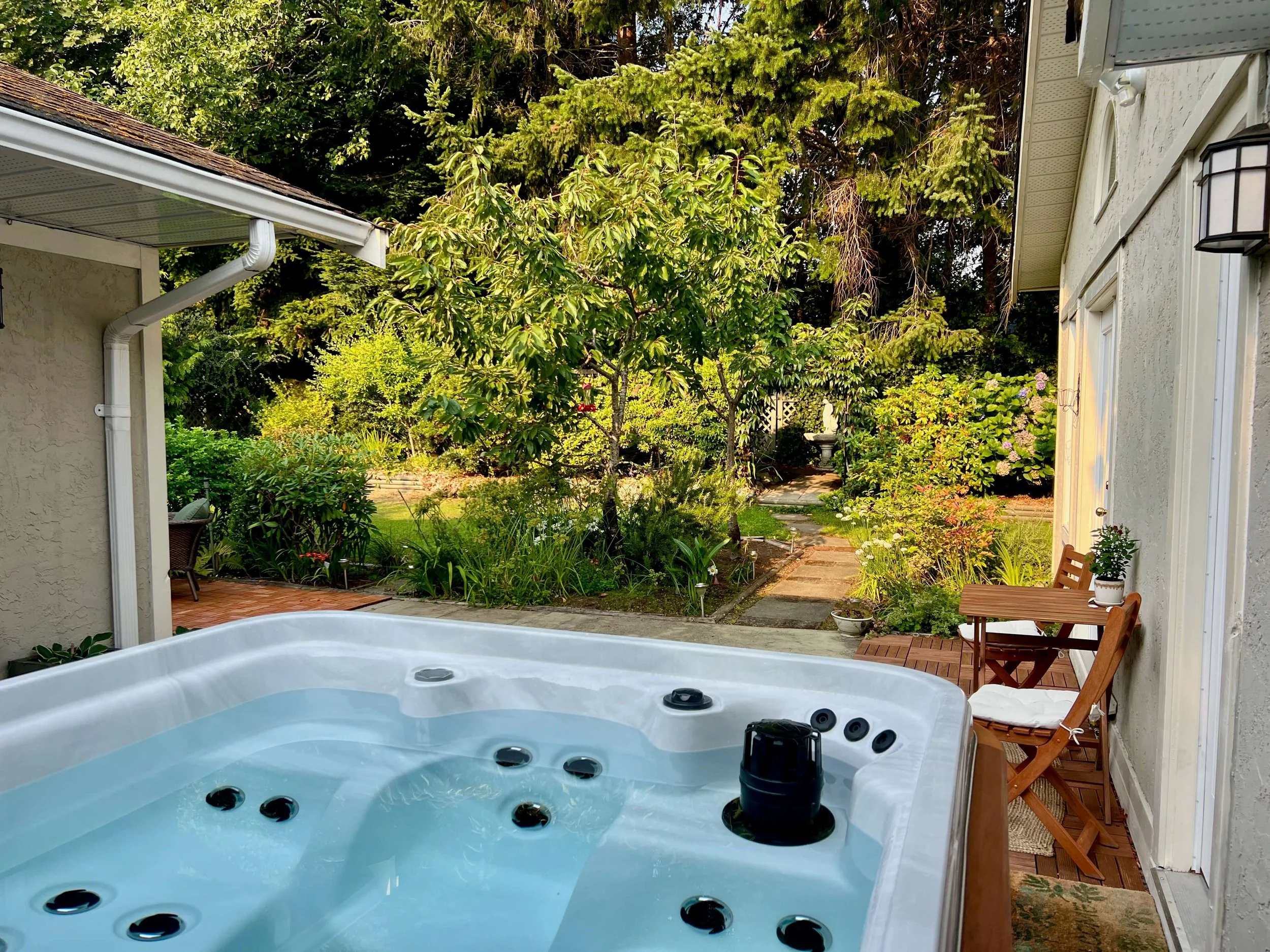 An outdoor hot tub with a wooden exterior, filled with water, on a tiled patio with a small wooden bench beside it, and a covered hot tub lid leaning against the wall.