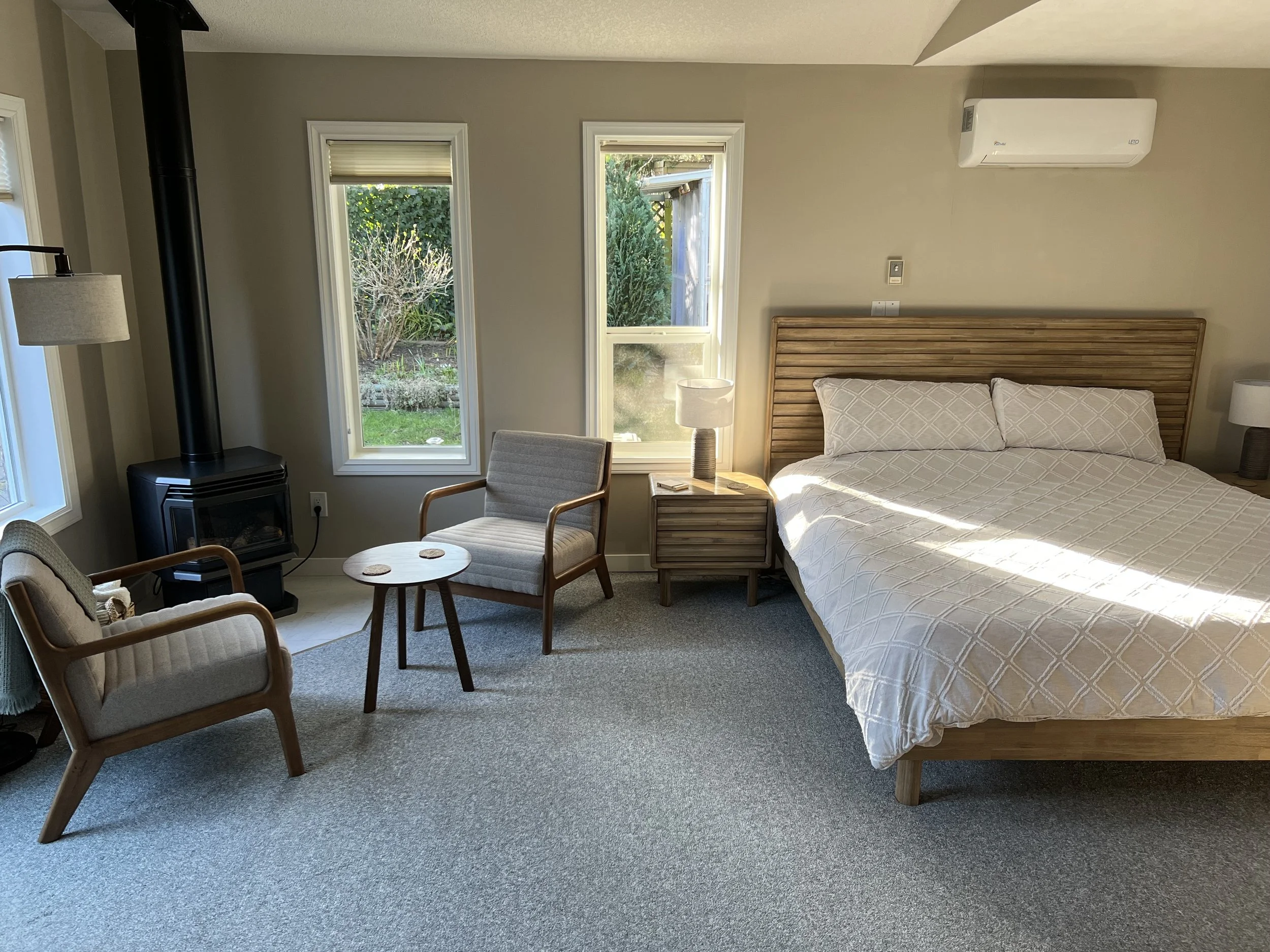 Cozy bedroom with a large bed, two side tables with lamps, two armchairs, a small round table with a book, a fireplace, a wall-mounted air conditioning unit, gray walls, carpeted floor, and three windows revealing a garden outside.