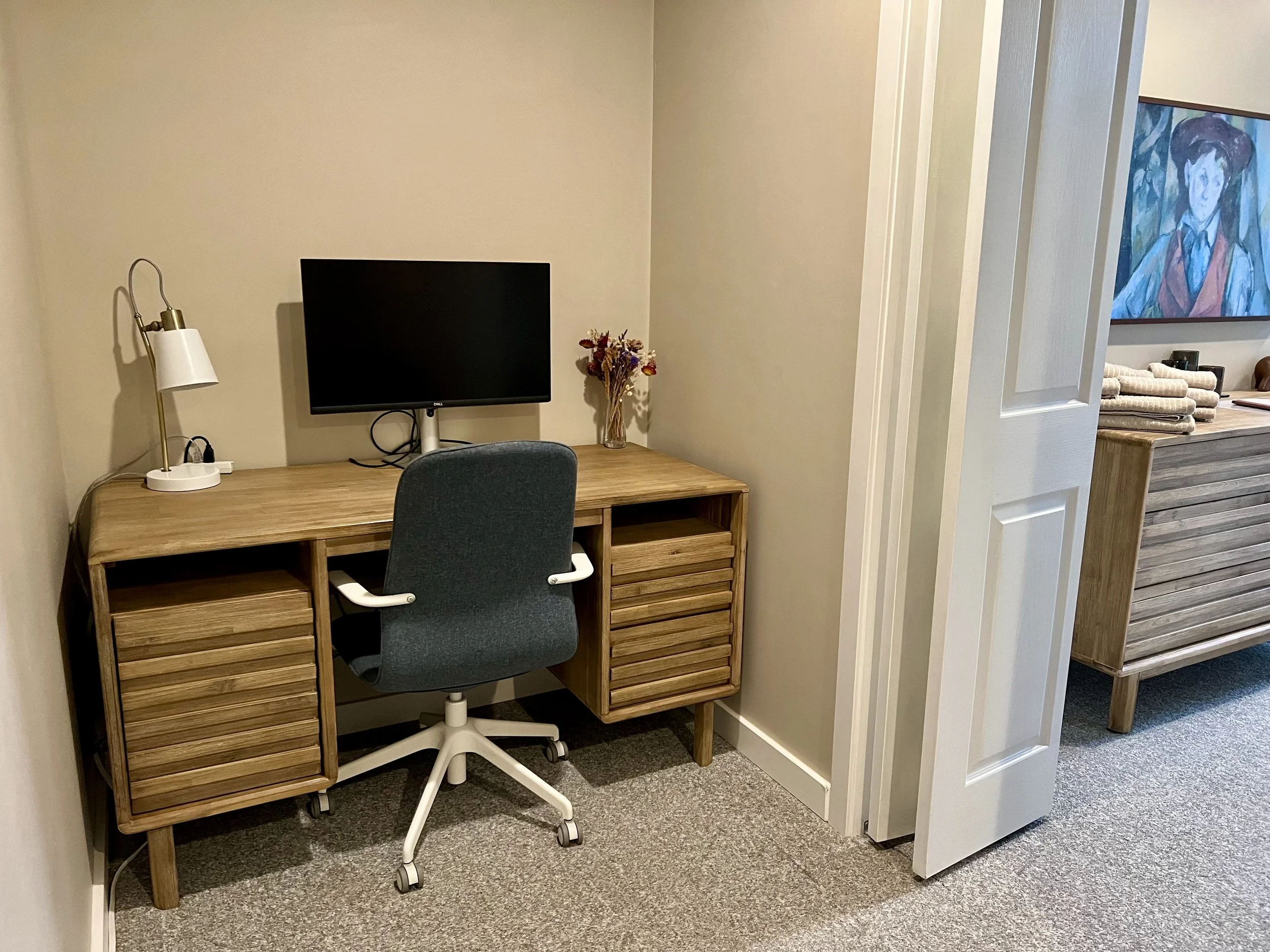 Wooden desk with a black monitor, white desk lamp, decorative dried flowers, and a grey office chair in a room.