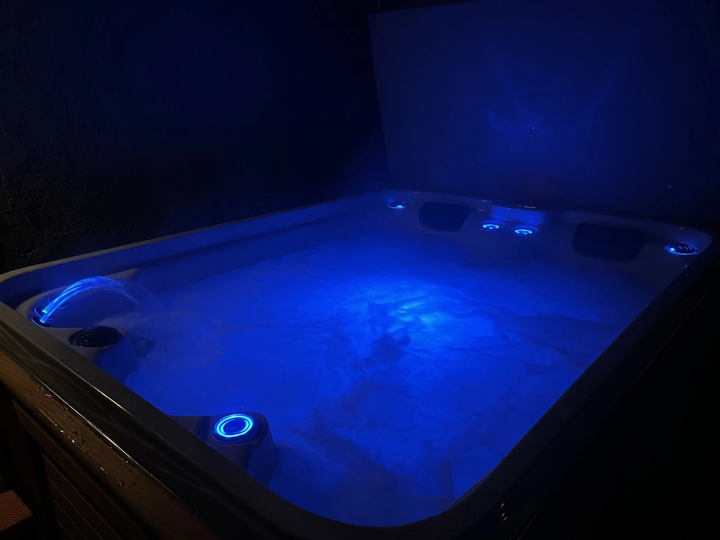 Outdoor hot tub illuminated with blue lights, surrounded by wooden decking and a wooden step, with the cover open, in an evening setting.