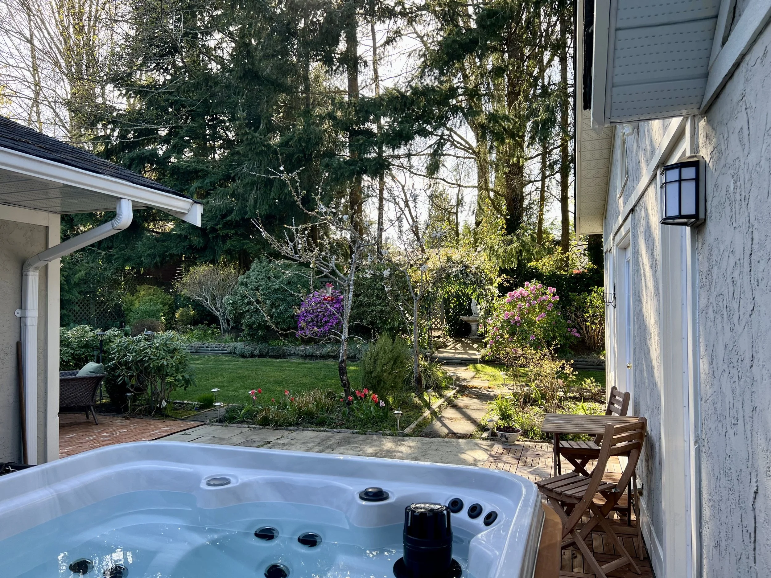 An outdoor hot tub with a wooden exterior, filled with water, on a tiled patio with a small wooden bench beside it, and a covered hot tub lid leaning against the wall.