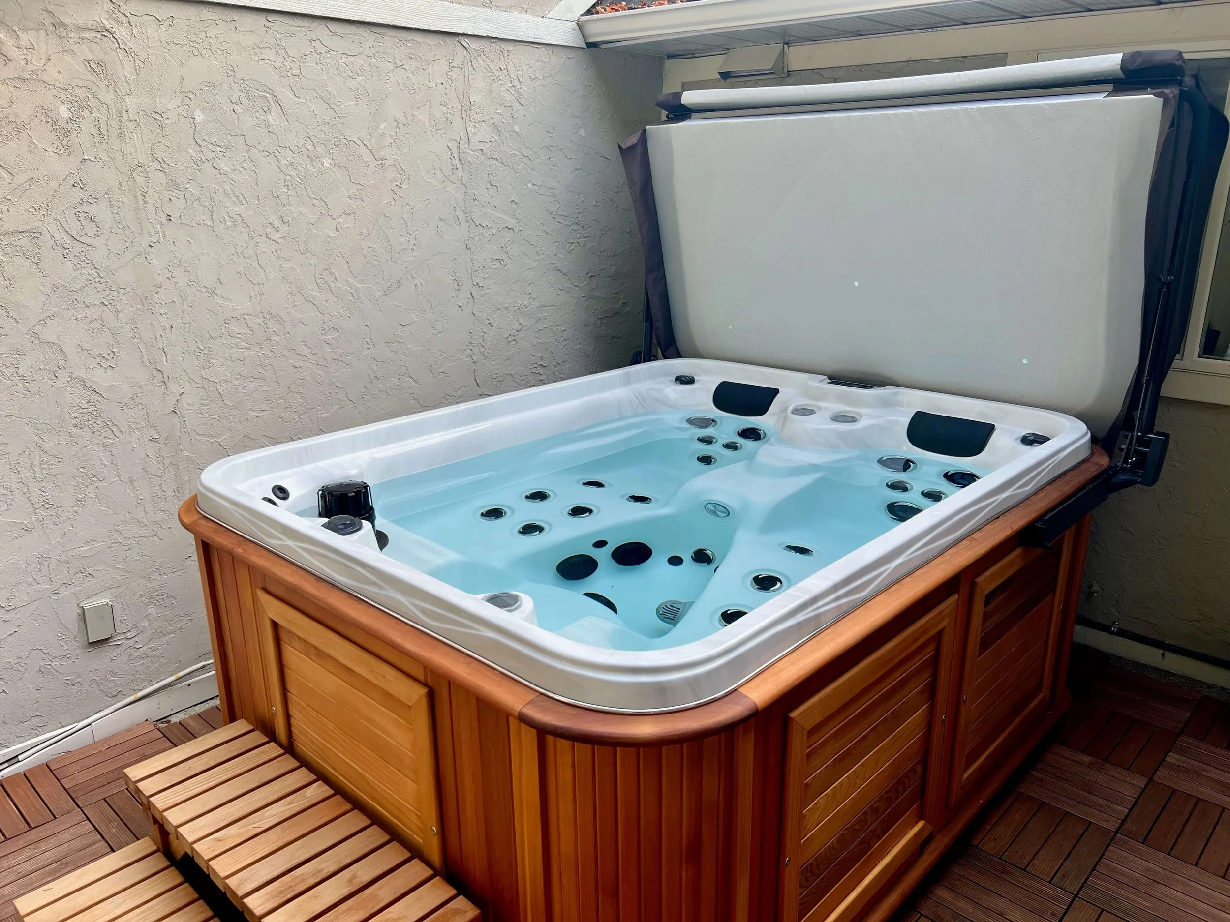 A hot tub with a wooden exterior and a partially open cover on a tiled patio, with a beige textured wall in the background.