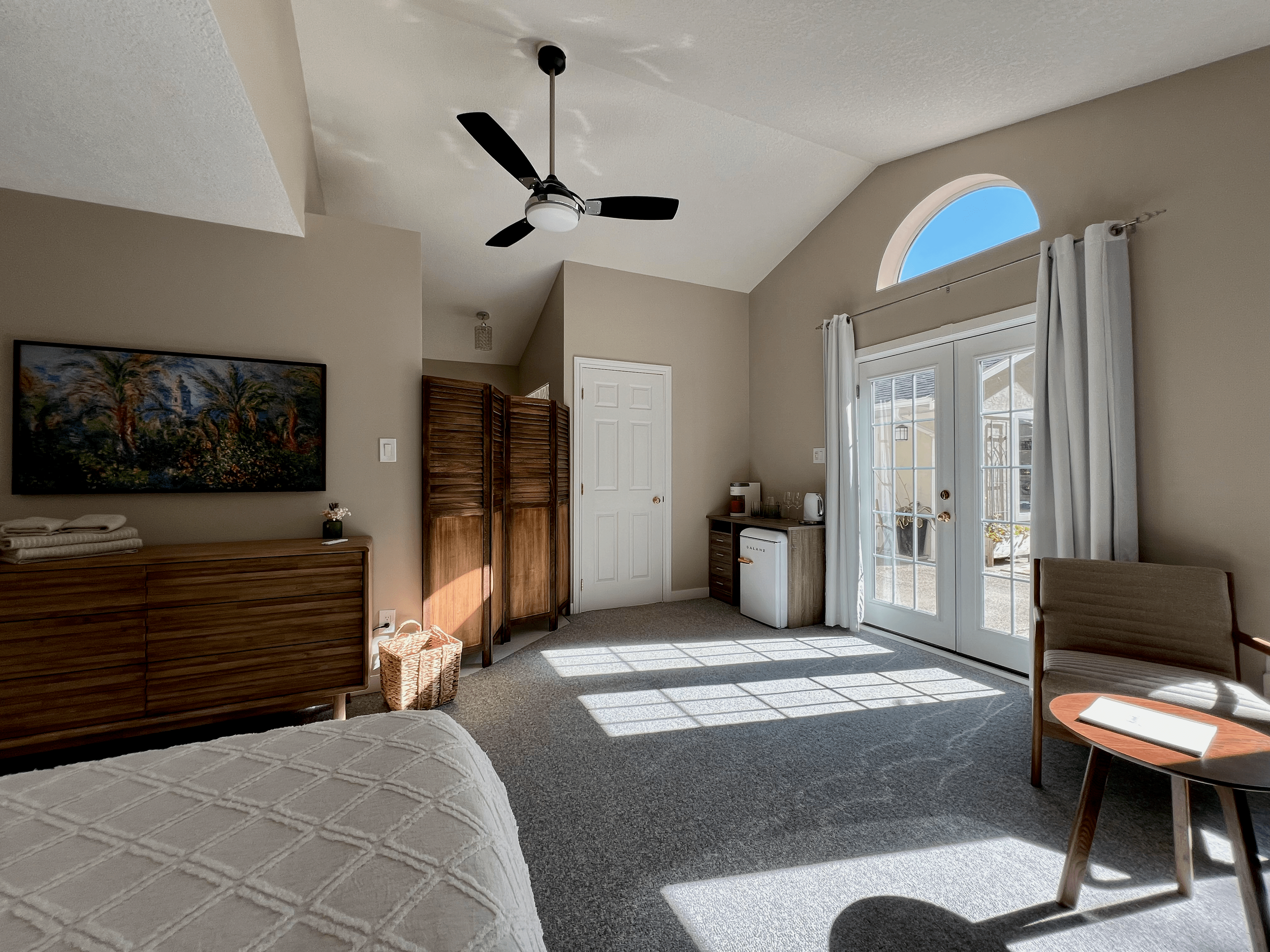 Bright hotel room with beige walls, a ceiling fan, a large window and glass doors leading outside, a wooden dresser, a small kitchenette area, and seating including a chair and side table with a laptop.