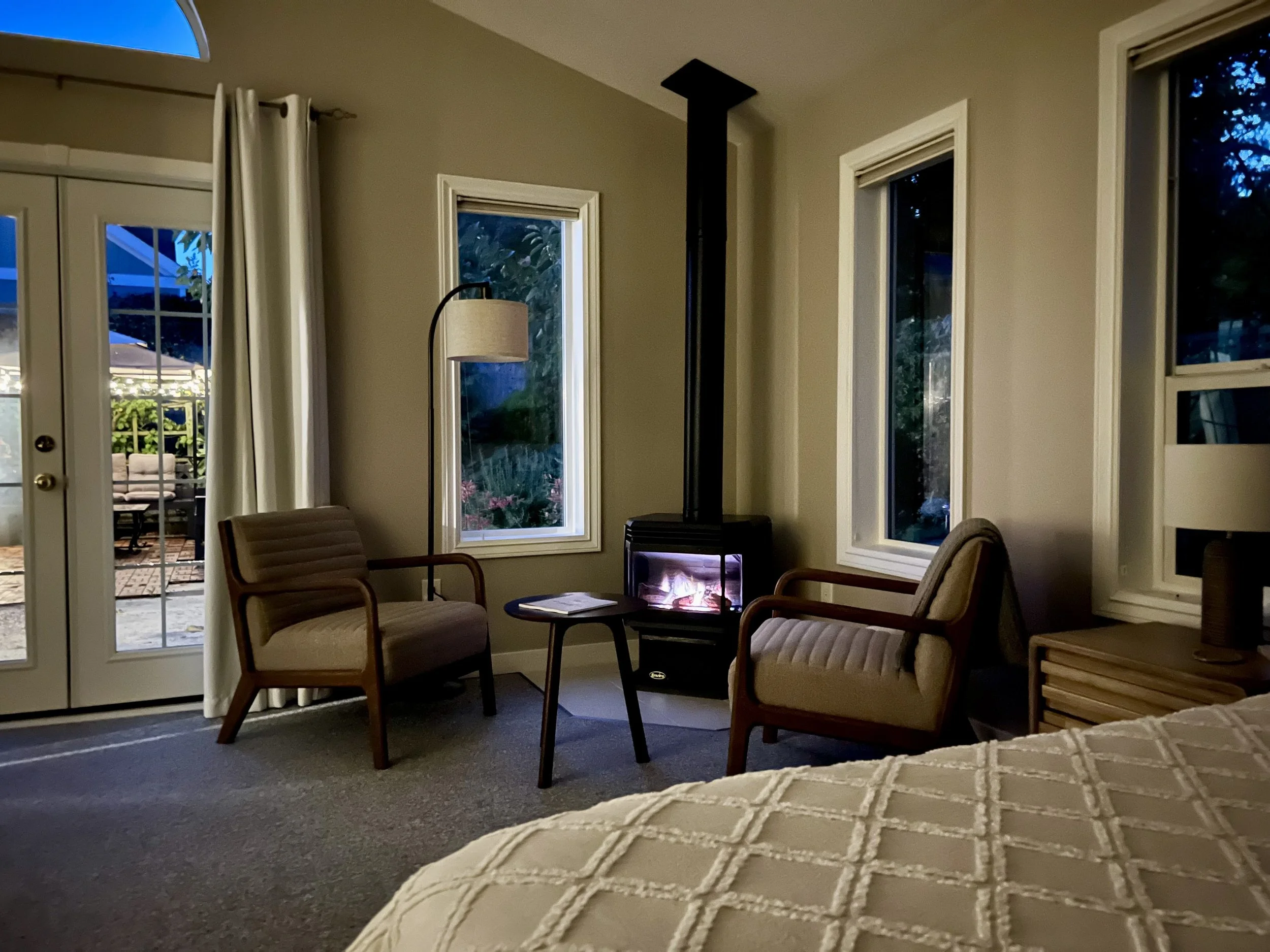 Living room with two beige armchairs, a small wooden table with magazines, a black wood stove with a tall chimney, a lamp with a beige shade, and three large windows showing outdoor greenery. There are French doors leading outside and part of a bed with a white quilted cover in the foreground.