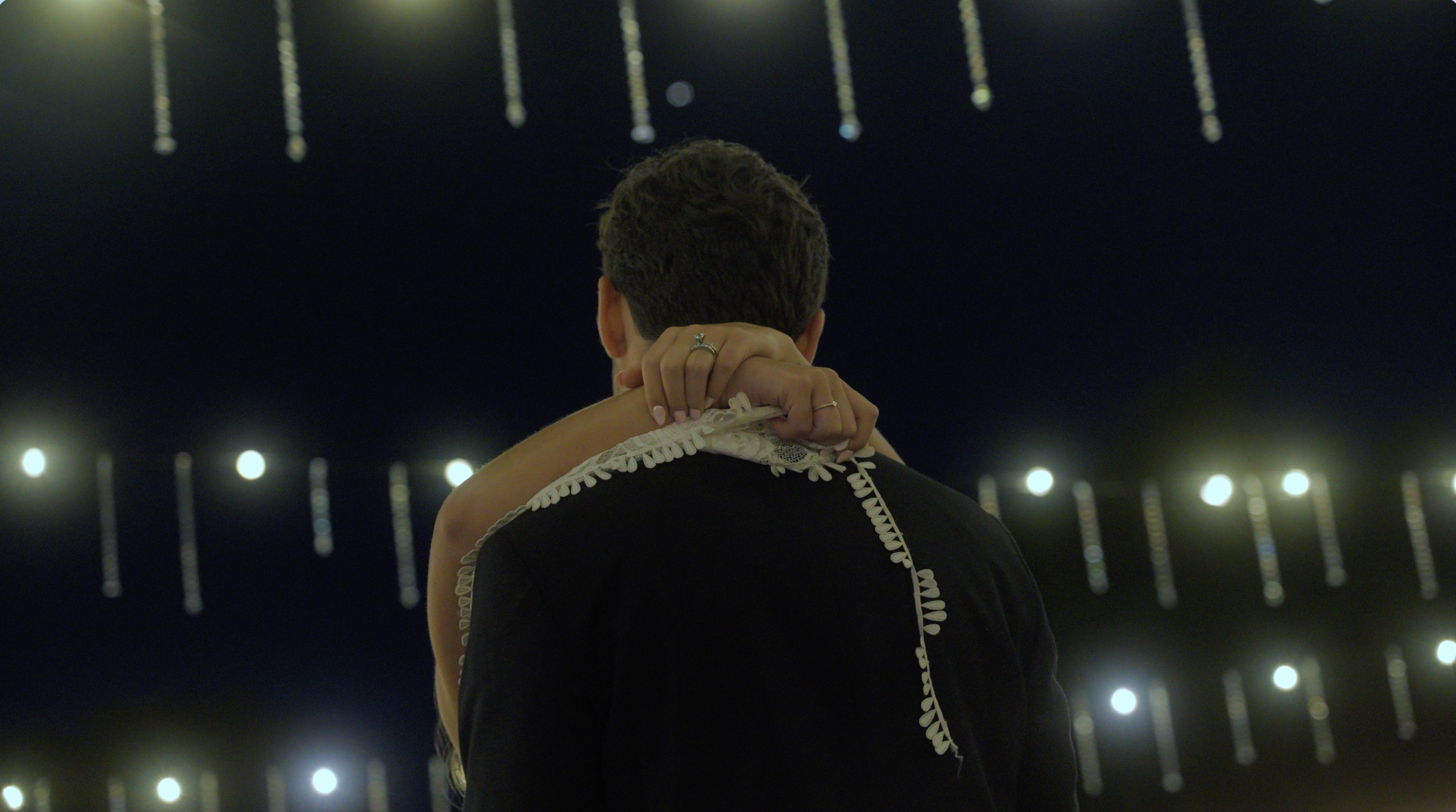 A woman is embracing a man from behind in a dark space illuminated by hanging lights or decorations in the background. The woman's arms are around the man's neck, and she is wearing rings on her fingers. The man is facing away from the camera, and their faces are not visible.
