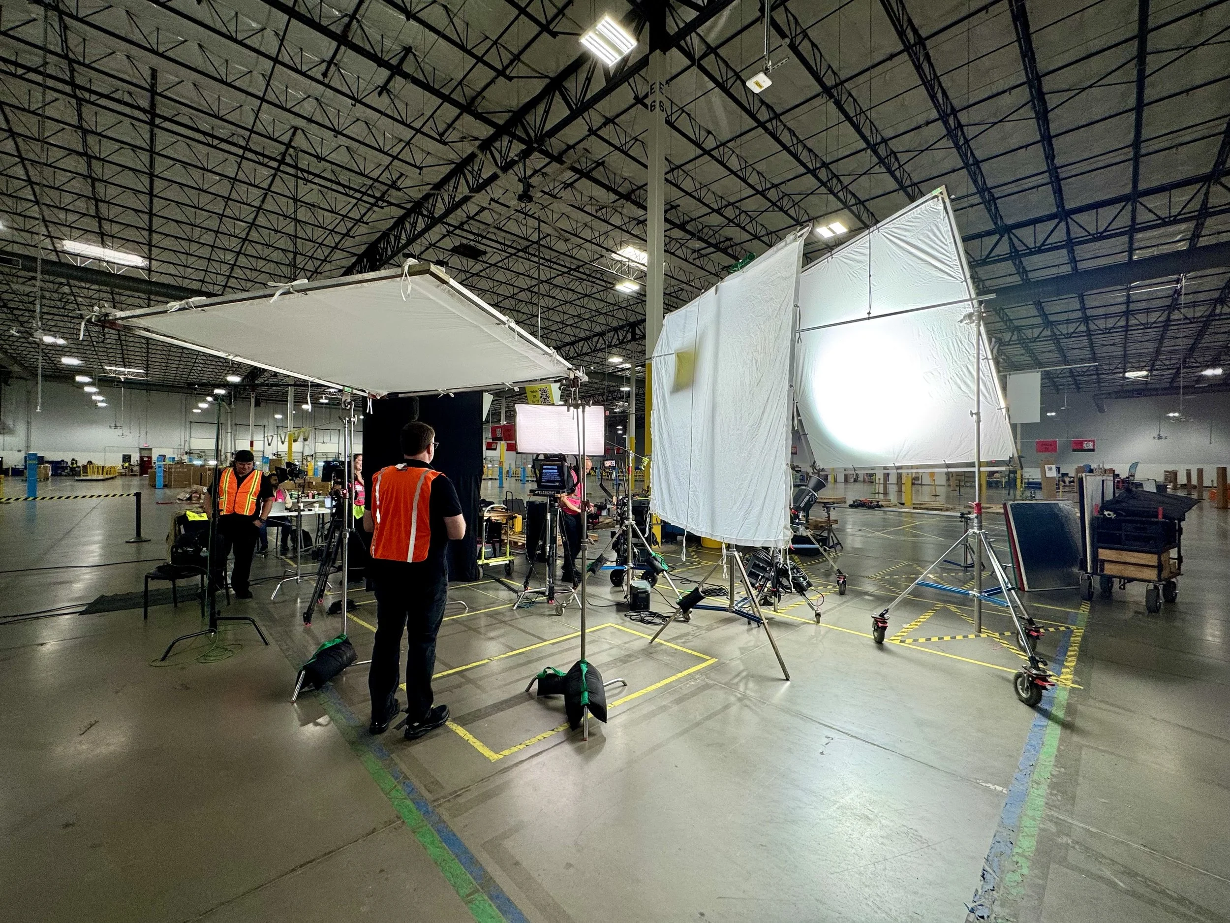 A professional video shoot setup in a large industrial warehouse with lighting and camera equipment, crew members wearing safety vests, and white fabric backdrops. San Antonio Grip Truck