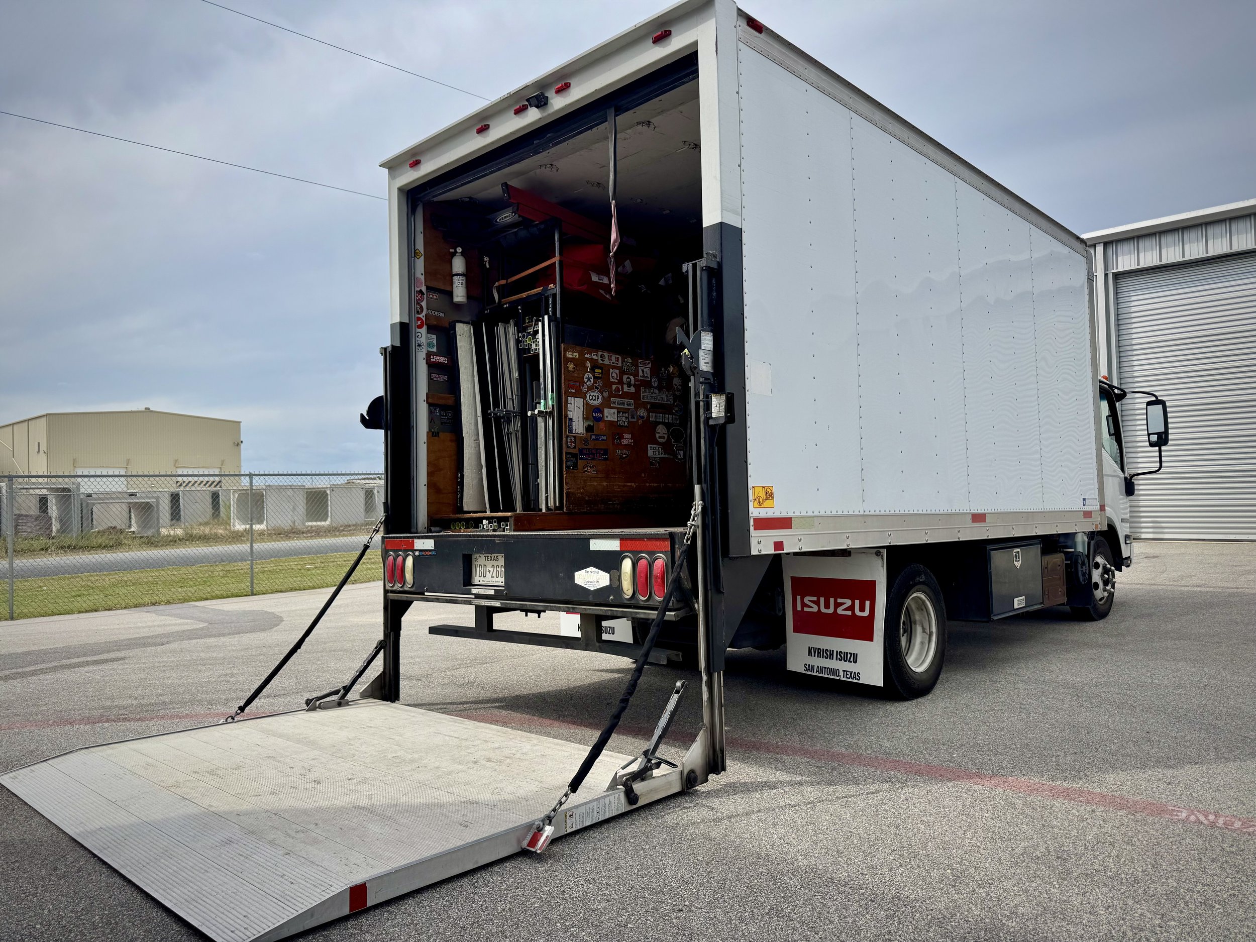 A white Isuzu moving truck with the back door open, showing a ramp extended and equipment inside on a cloudy day. San Antonio Grip Truck