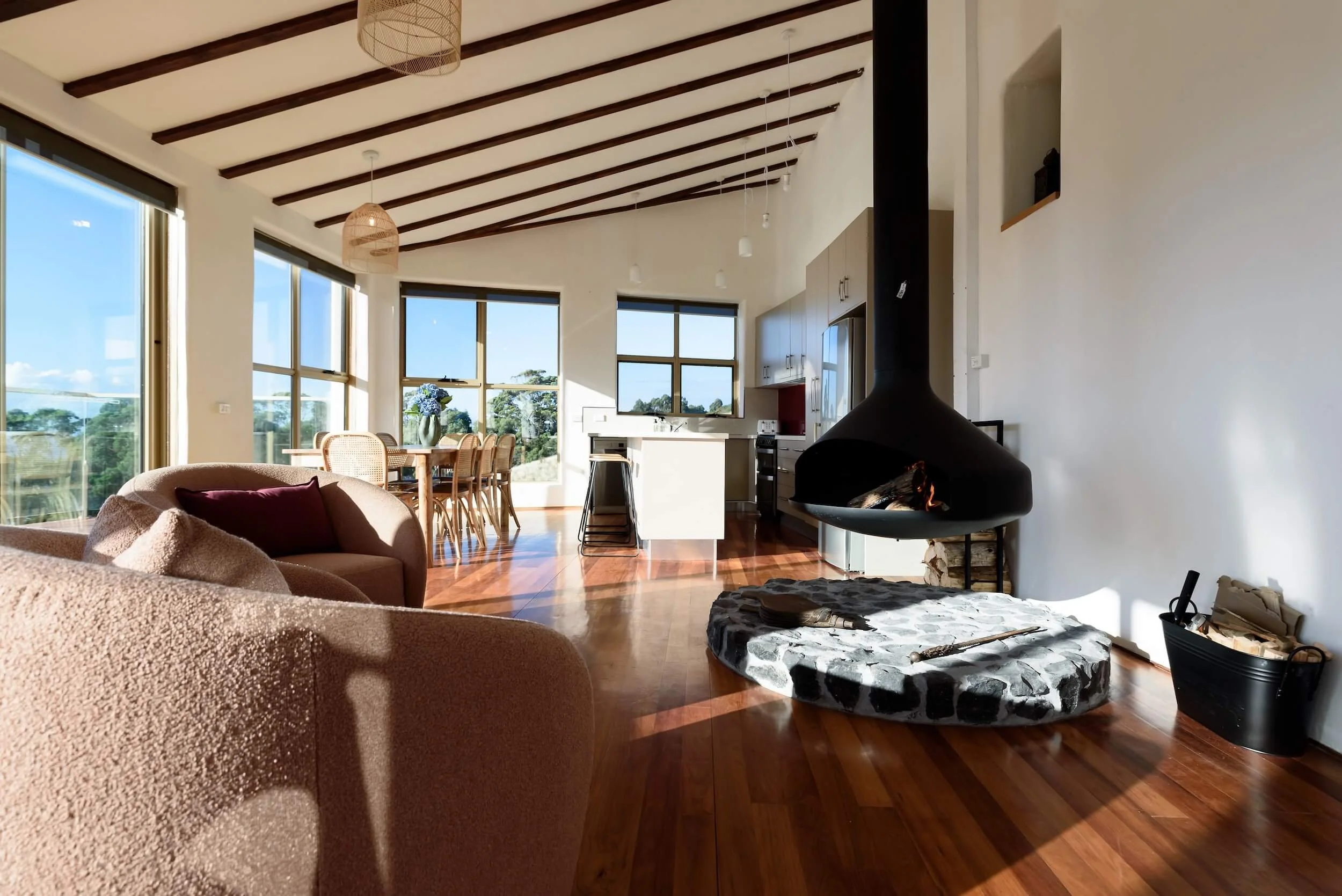 Living room with large windows, wood floor, beige sofa, black fireplace, and dining table with chairs near the windows.