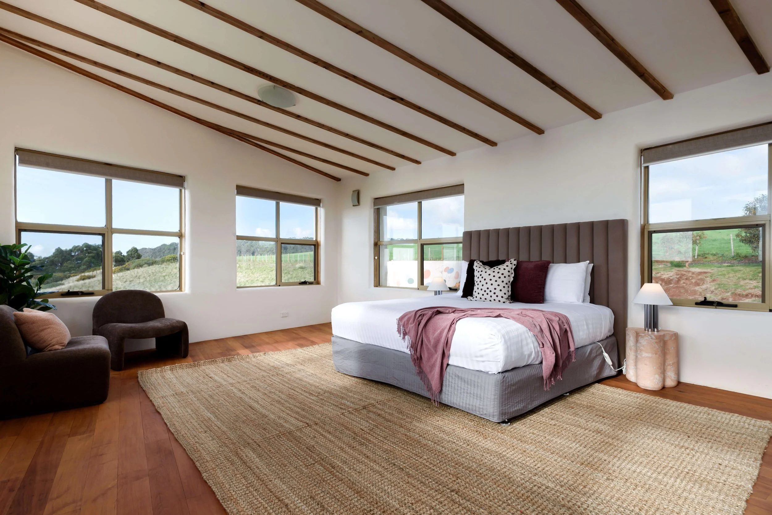 A spacious bedroom with a modern design, featuring a white bed with pillows and a blanket, a beige tufted headboard, large windows with views of greenery, a brown armchair with a pillow in the corner, a woven rug on wooden flooring, and natural light filling the room.