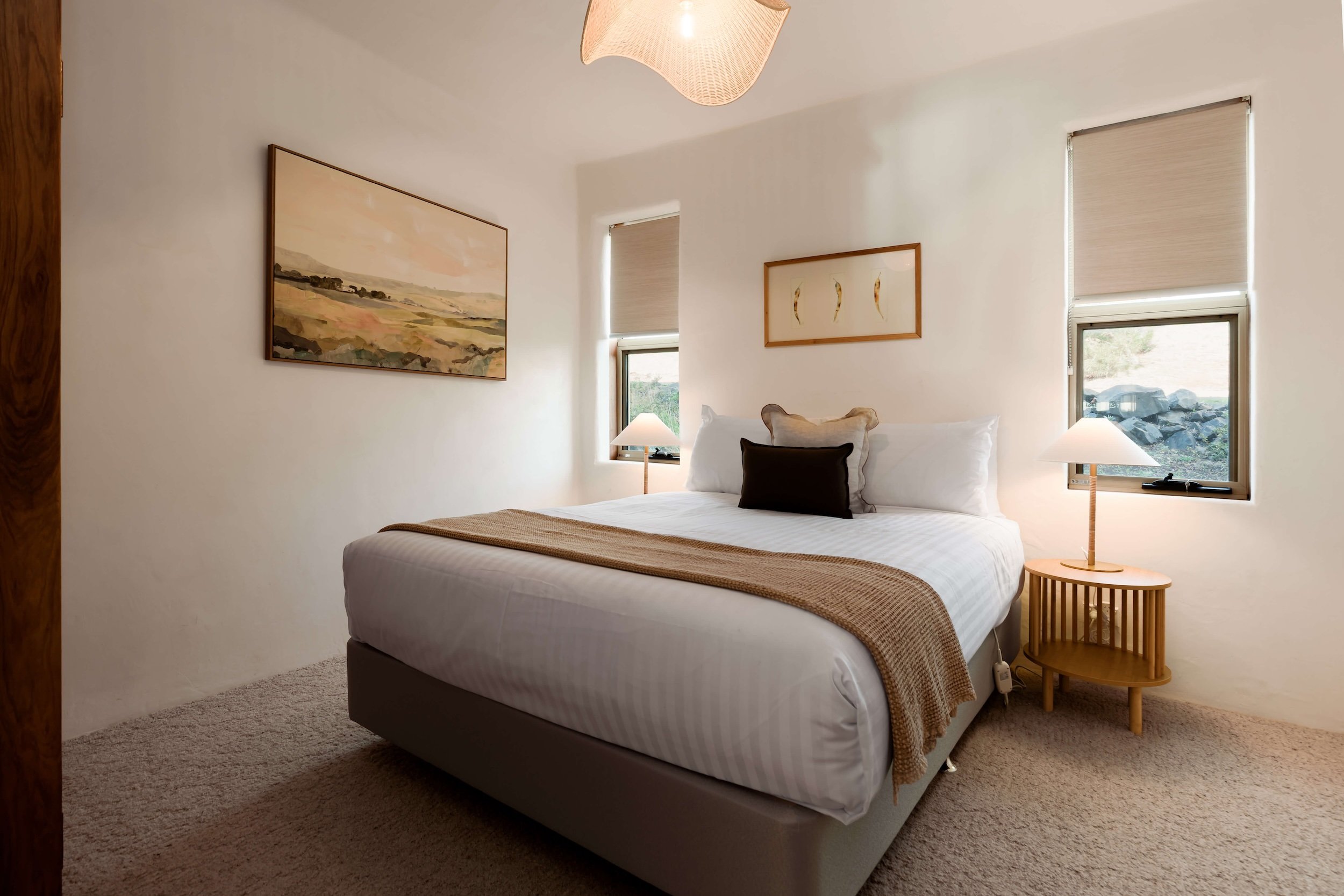 A bedroom with a large bed covered in white linens and a beige throw blanket, two pillows, and a small dark pillow. There are three windows with beige roller shades, a wooden nightstand with a lamp, and two framed artworks on the walls. The room has beige carpet and a white ceiling with a modern pendant light.