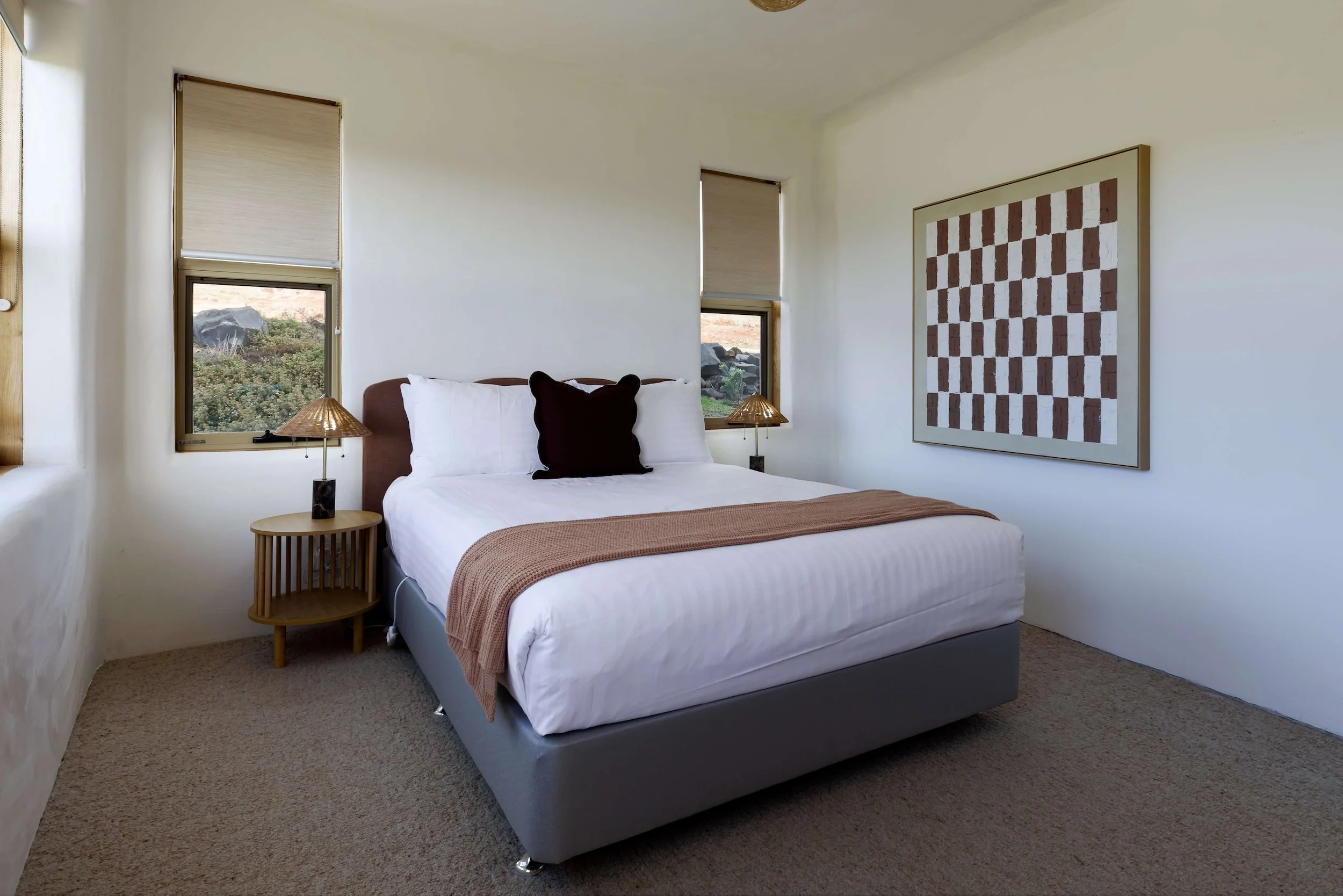 A bedroom with a bed, window views of nature, framed artwork, bedside lamps, and beige carpeting.