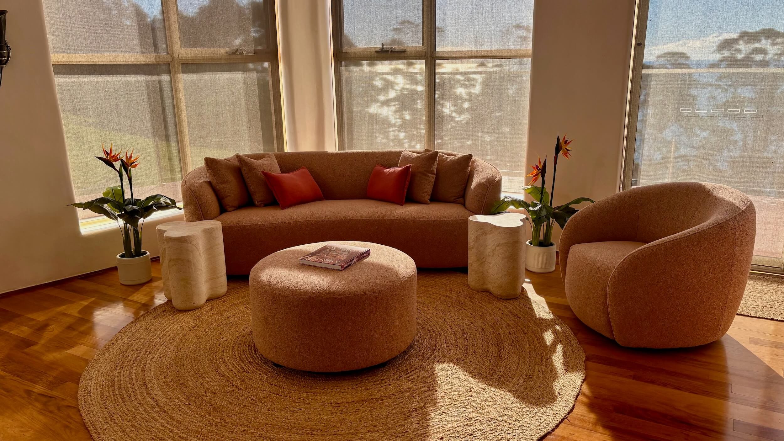 Sunlit living room with two chairs, a sofa with red pillows, a round ottoman with a book, two potted plants, and large windows with sheer curtains.