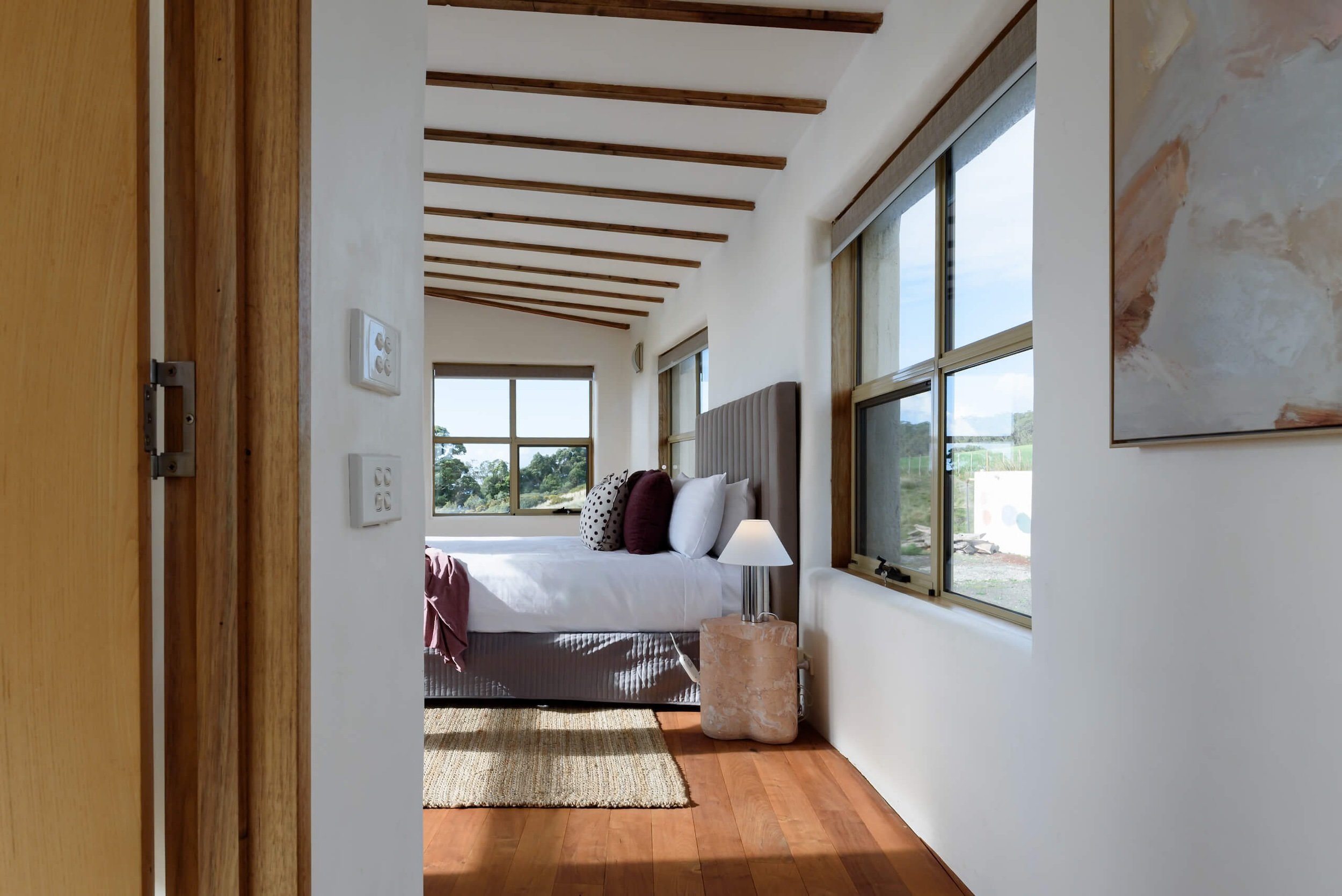 A bedroom with a bed, pillows, a bedside table with a lamp, large windows showing trees outside, and a ceiling with wooden beams.