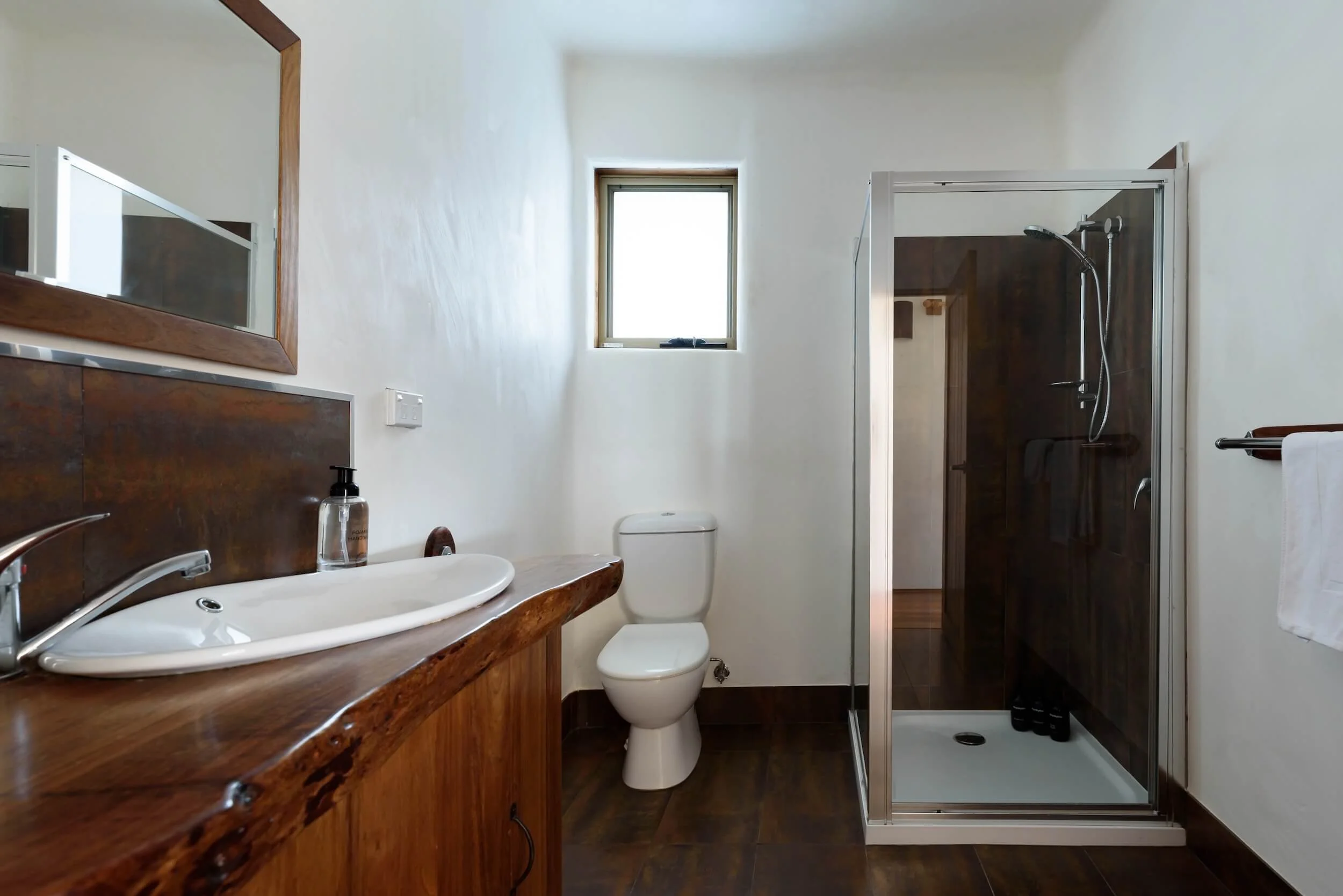 A small bathroom with a wooden vanity sink, a toilet, a small window, and a glass shower enclosure in the corner.