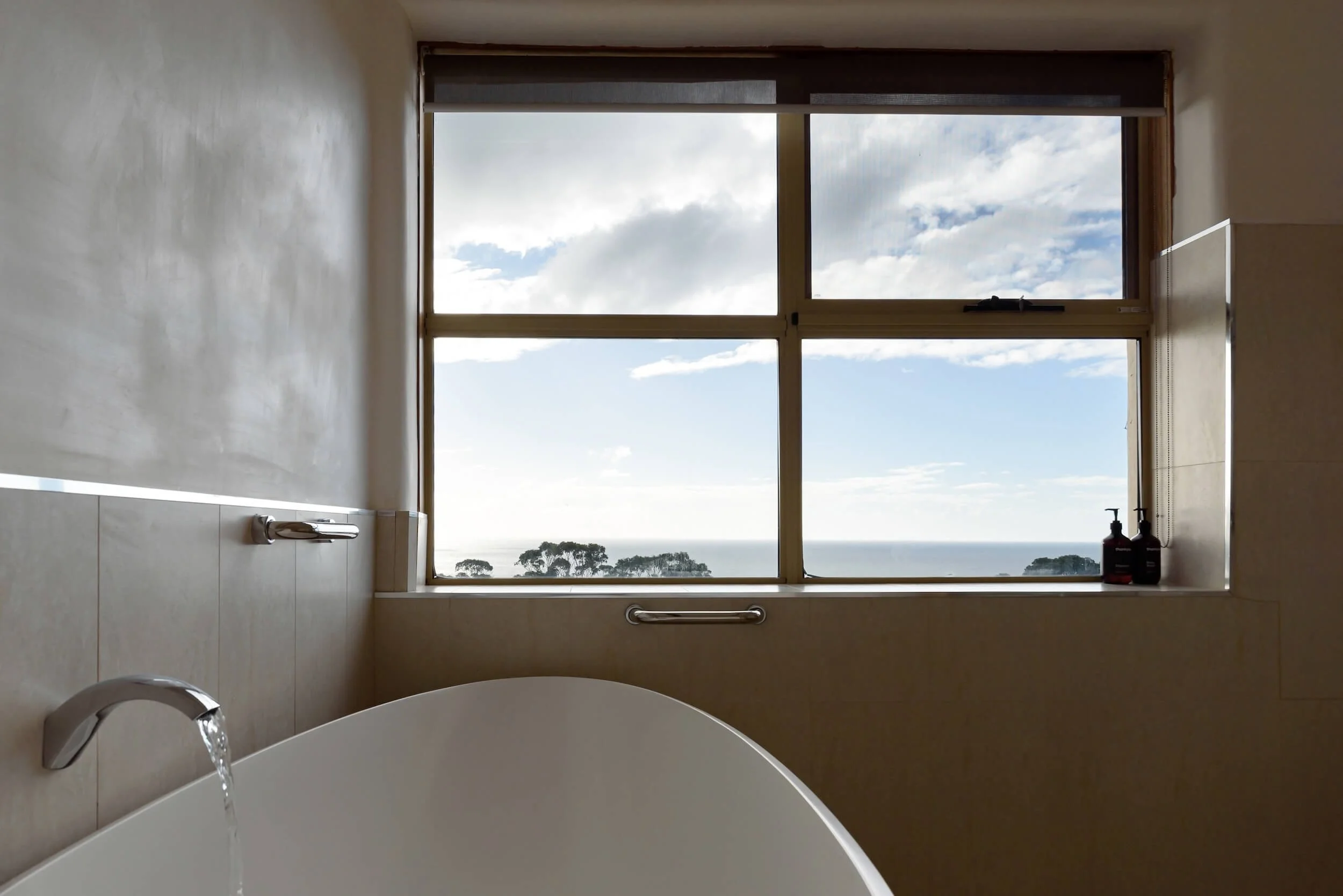 Bathroom with a large window showing a cloudy sky and distant trees, a bathtub with water flowing from a faucet, and two bottles on the windowsill.