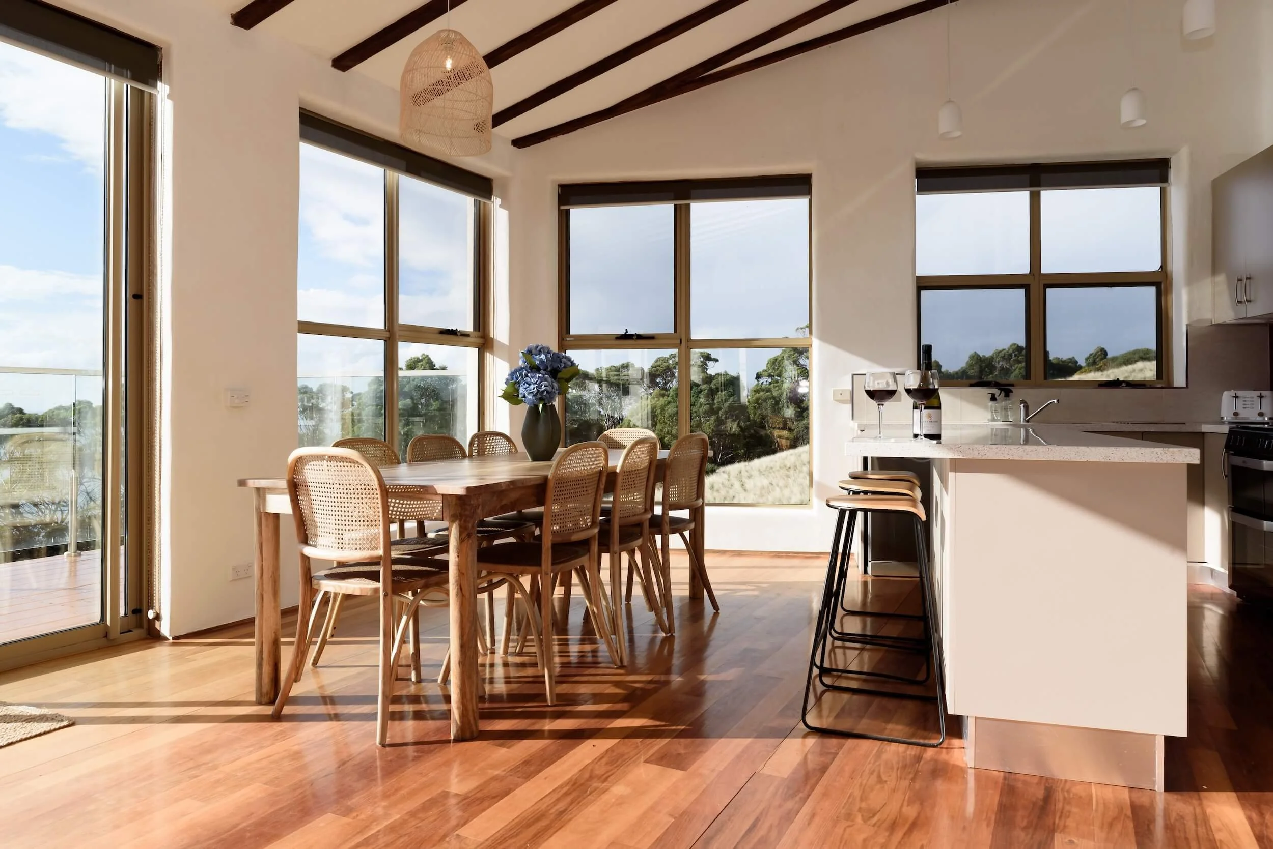 Bright dining area with large windows, wooden dining table with chairs, vase with blue flowers, and a kitchen counter with wine glasses and a bottle.