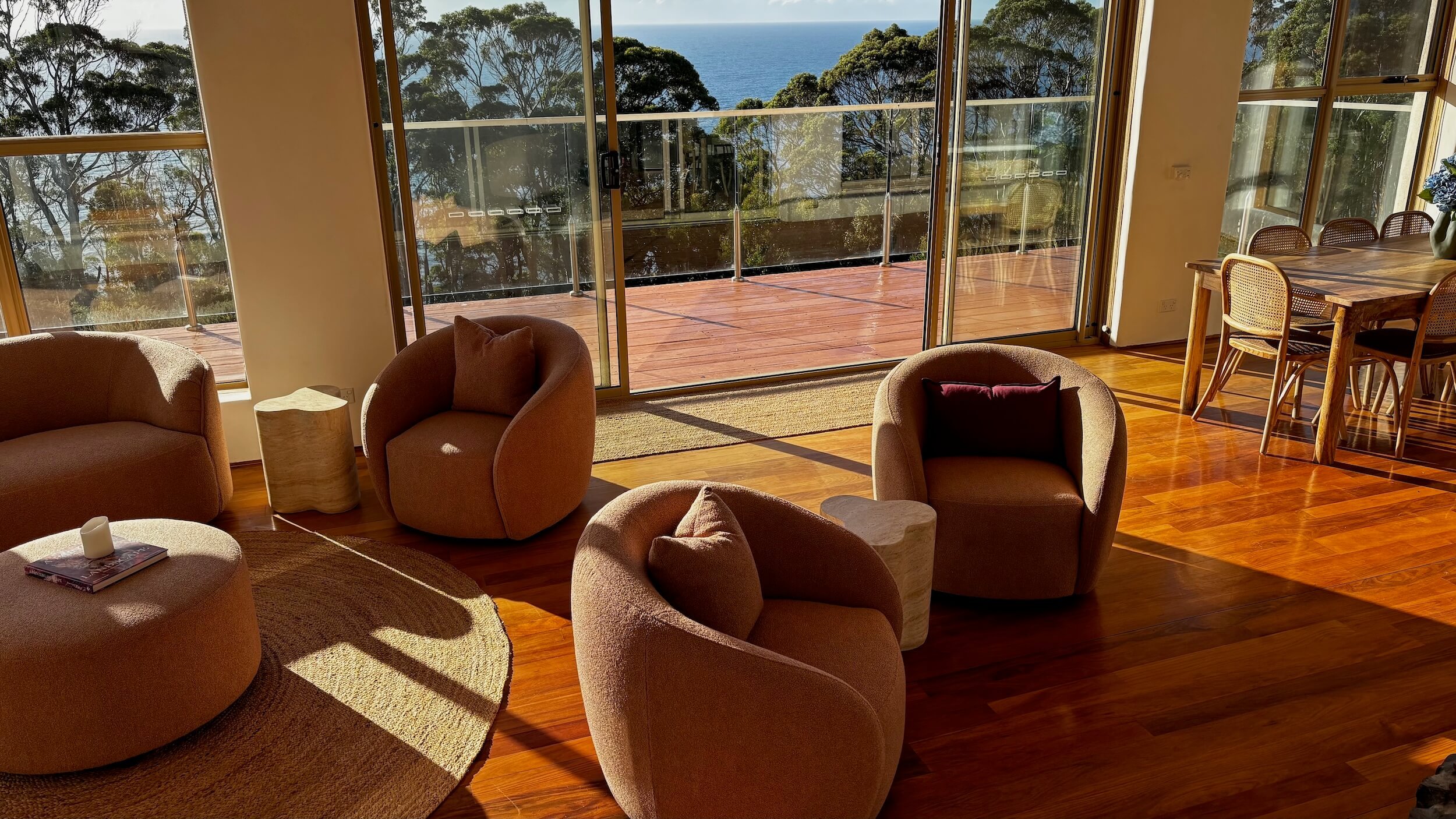 Living room with sliding glass doors opening to a balcony overlooking the ocean, with trees visible in the background. The room features a beige sofa and armchairs, a round woven rug, and a wooden dining table with chairs, illuminated by natural sunlight.