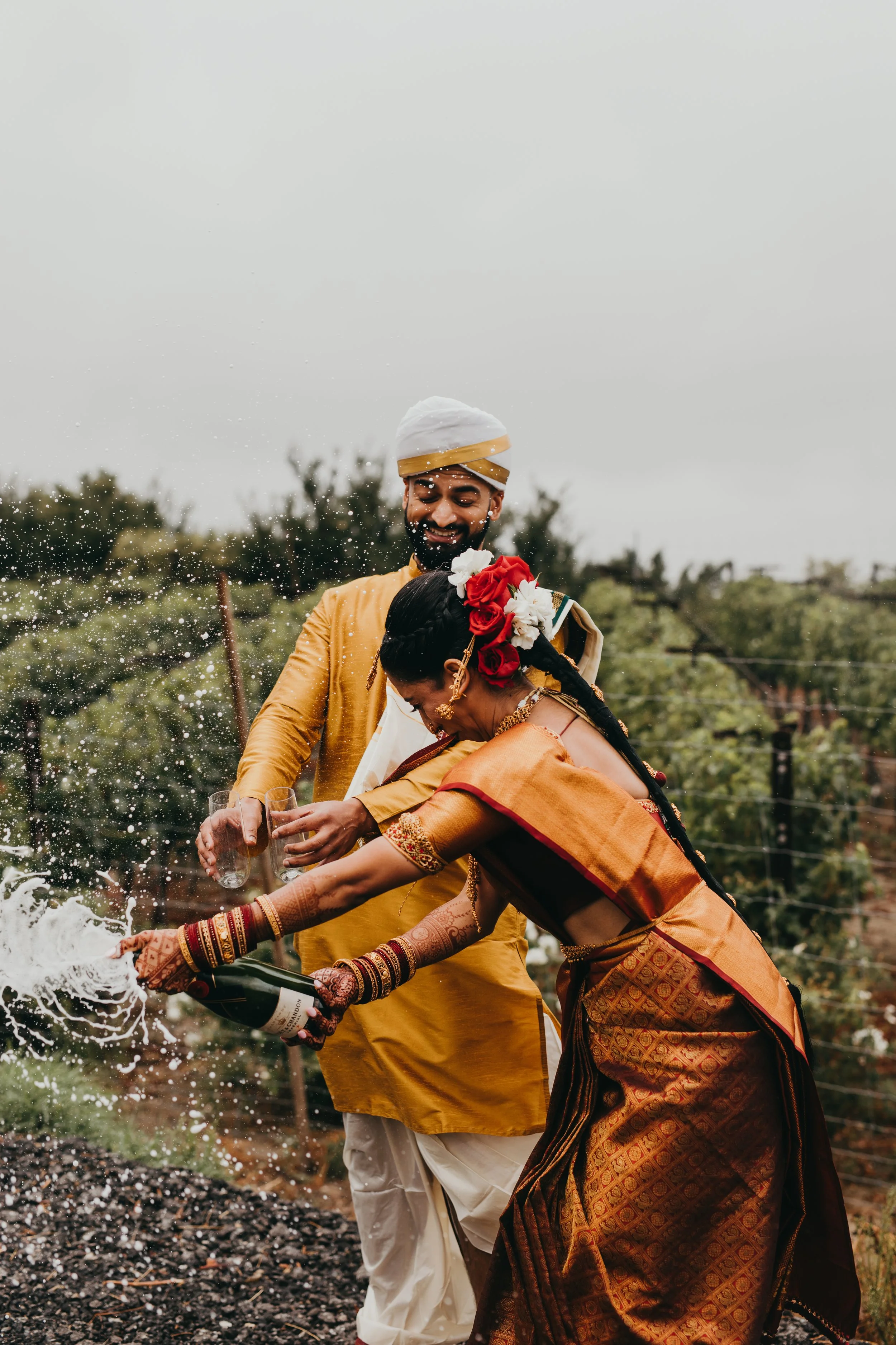 Indian couple in traditional attire celebrating their wedding outdoors with champagne, water splashing as they celebrate.