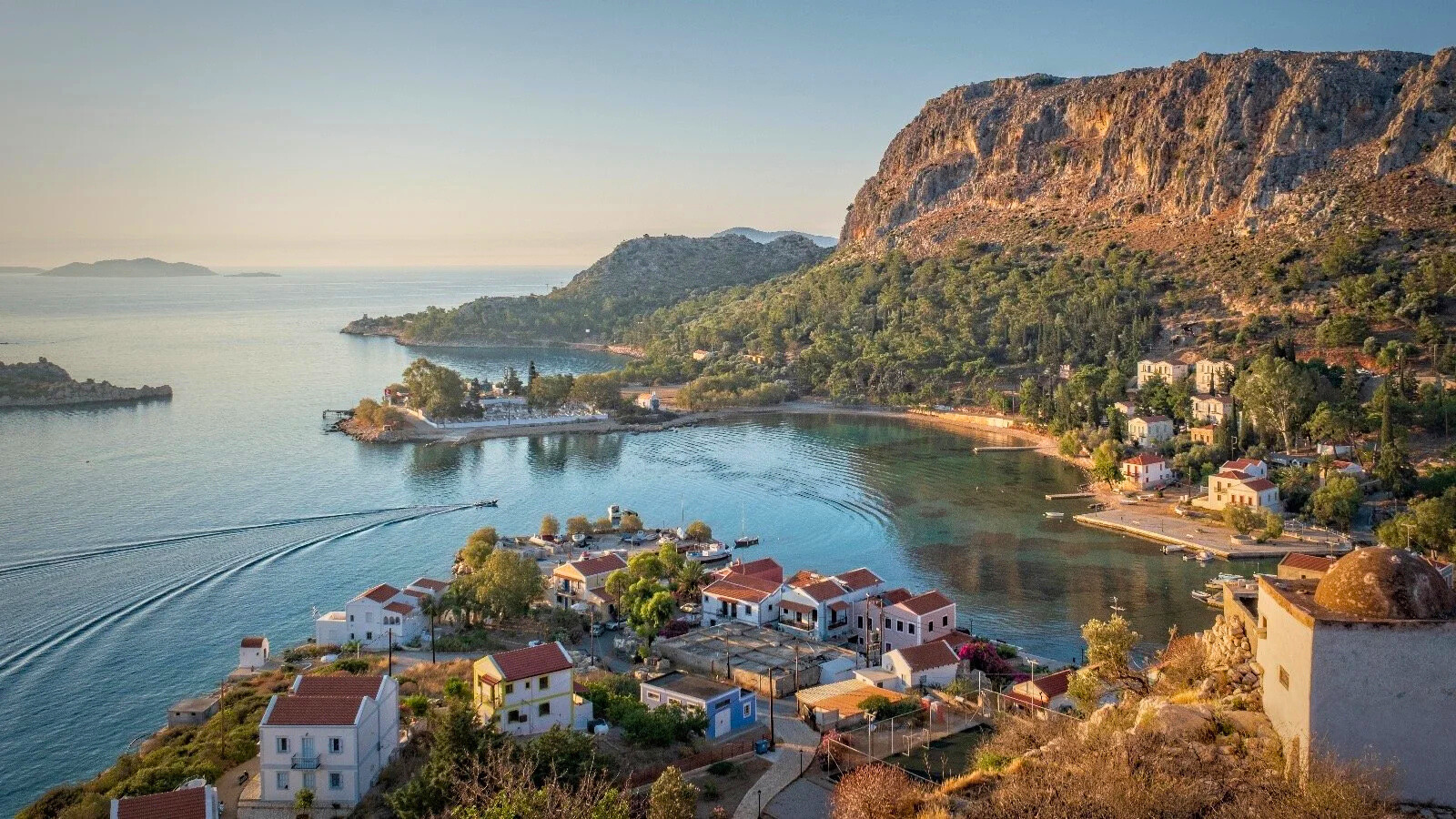 Coastal village with white buildings, boats on calm water, green hills, and rocky cliffs during sunset.