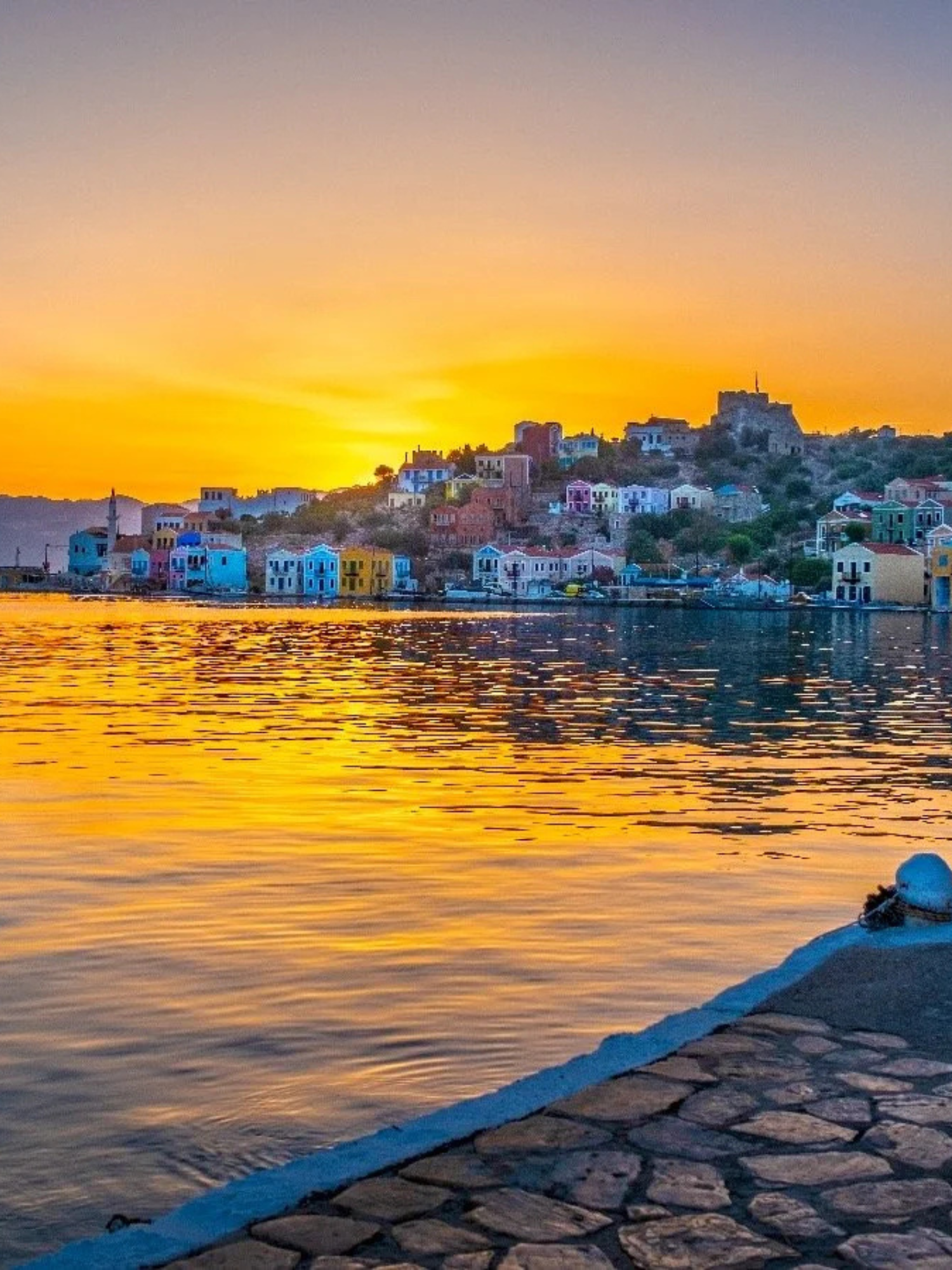 Colorful houses along a hillside by a body of water at sunset, with calm water reflecting the sky.