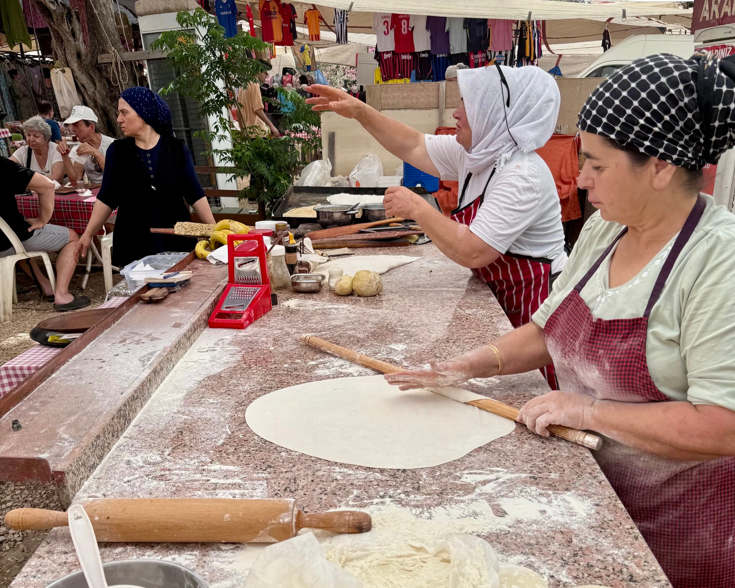 Two women in headscarves and aprons preparing dough on a large kitchen counter at an outdoor market or festival, with people in the background and hanging clothes above.