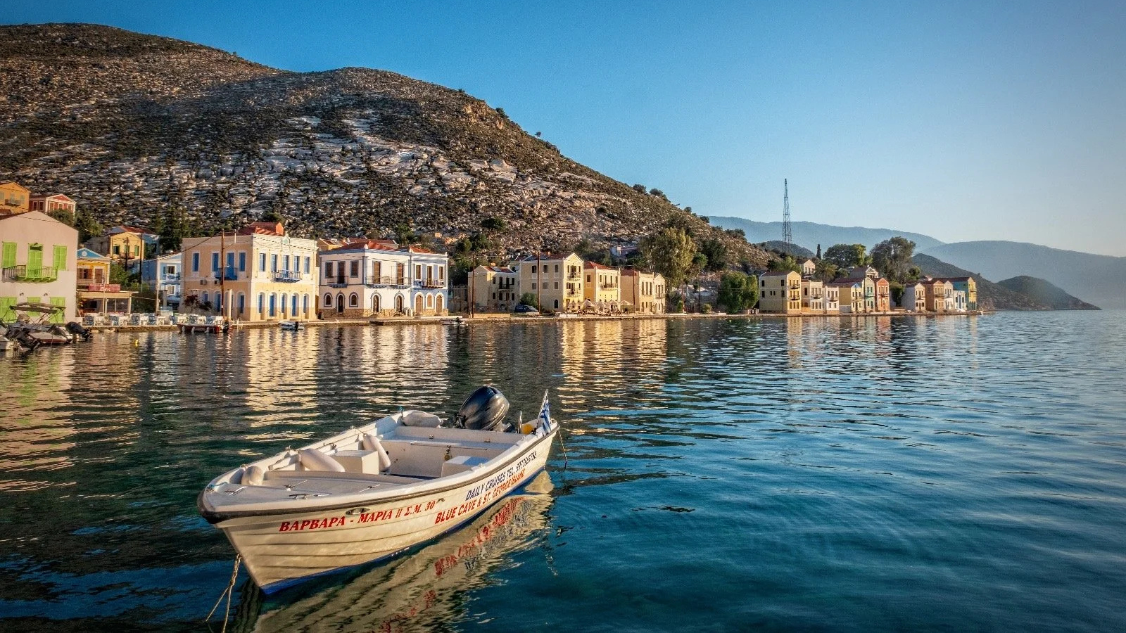 A boat floating on calm water with colorful houses along the shoreline and a mountain in the background.