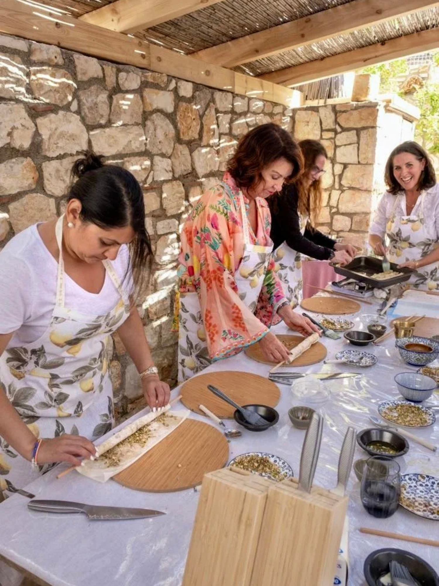 Four women preparing and cooking food outdoors under a wooden shelter, using rolling pins and various bowls and ingredients on a long table.