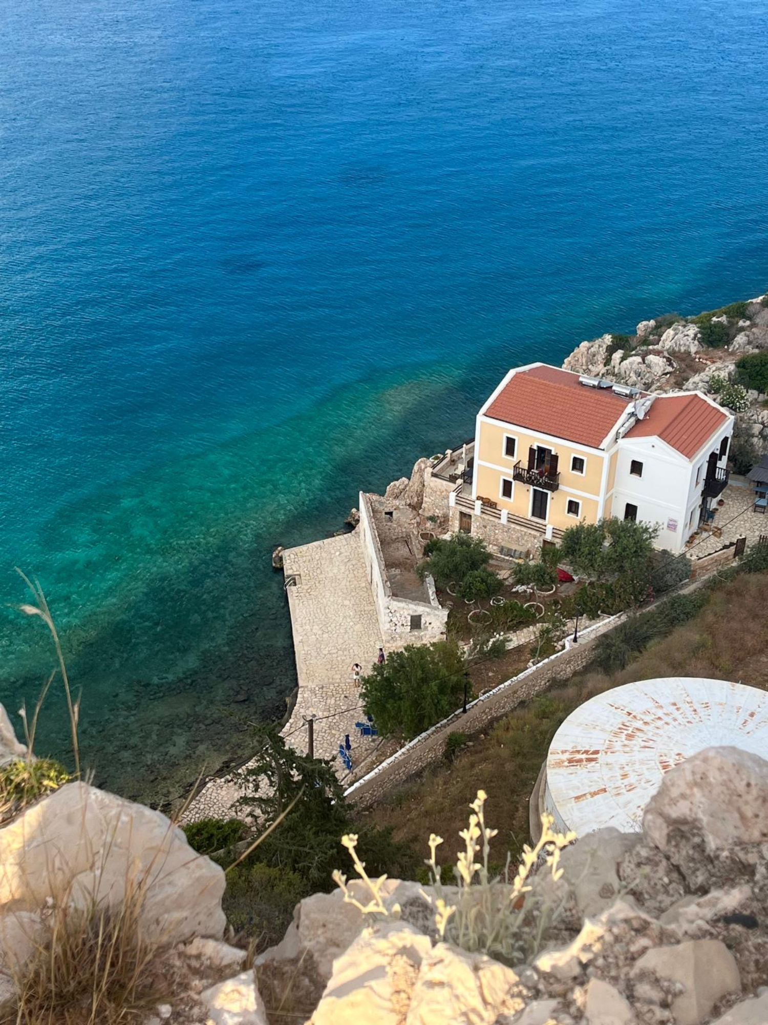 An aerial view of a house with a red roof near the coast, overlooking clear blue and green water, with rocky terrain and small trees nearby.