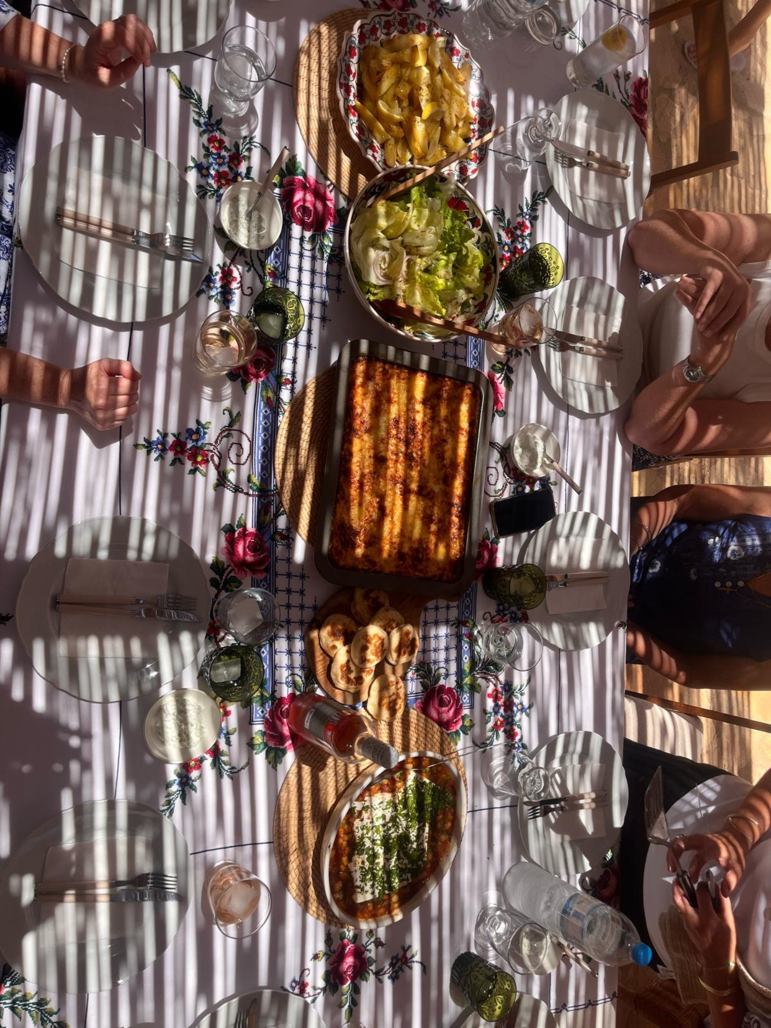 A table set with various dishes including a baked casserole, a bowl of salad, roasted potatoes, flatbreads, and condiments, with people around it during daytime.