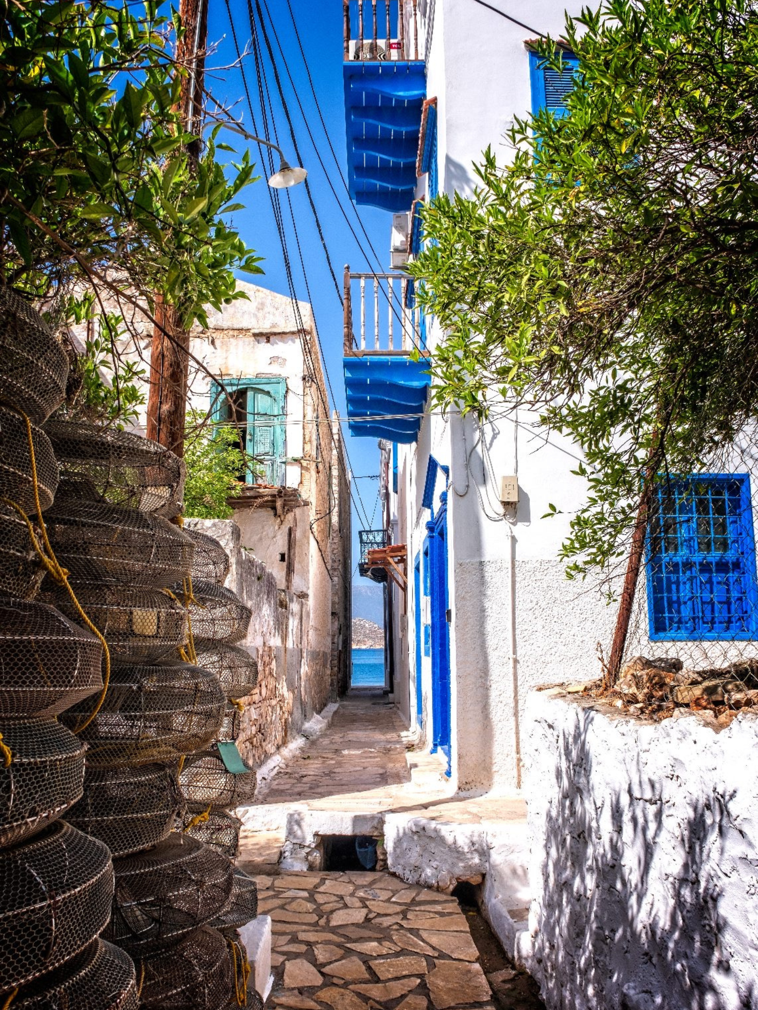 Narrow stone alleyway in a Mediterranean village with white buildings, blue shutters, and laundry hanging, under a clear blue sky.