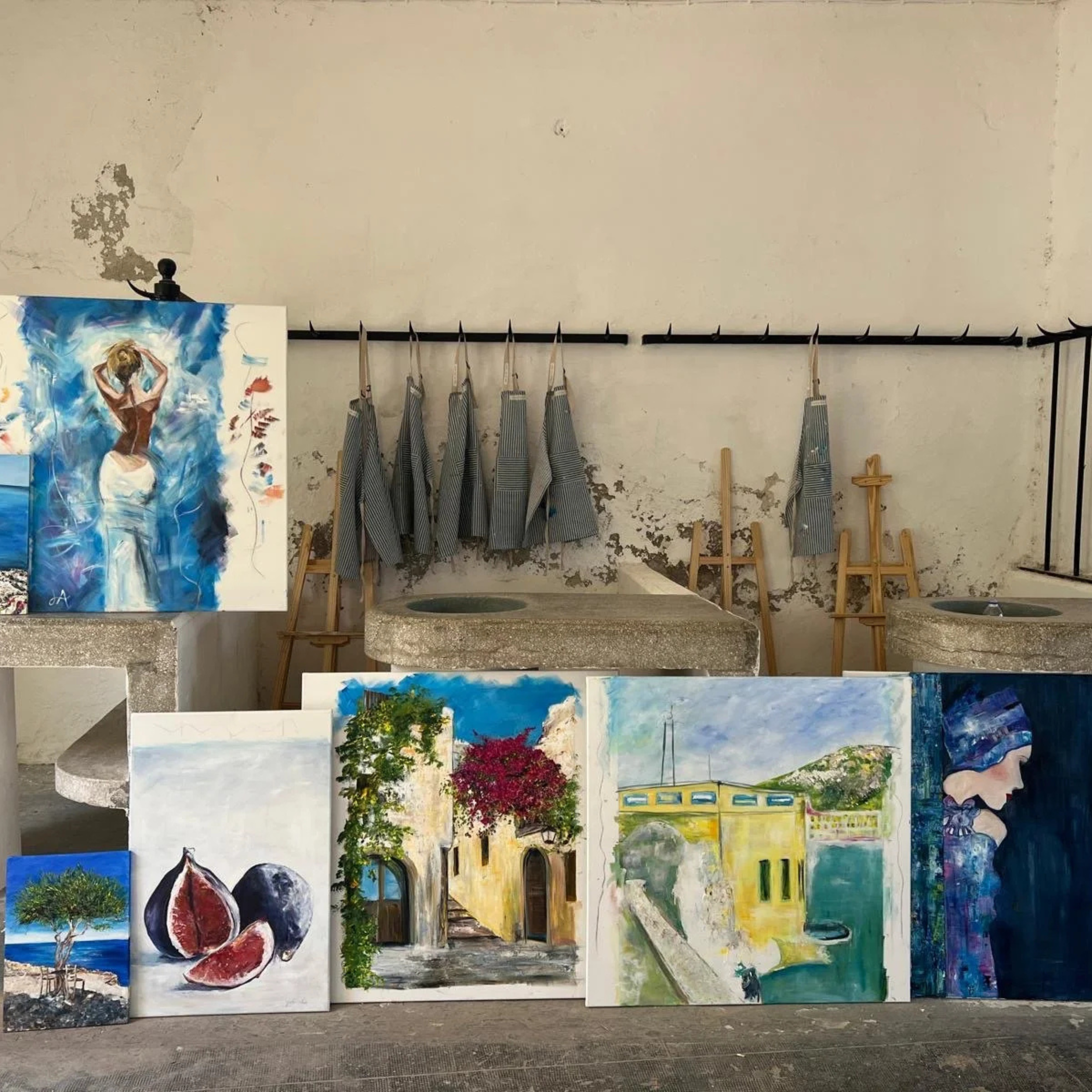 Various paintings displayed on the floor and a painting on an easel; aprons hanging on a rack in a rustic art studio with worn walls.