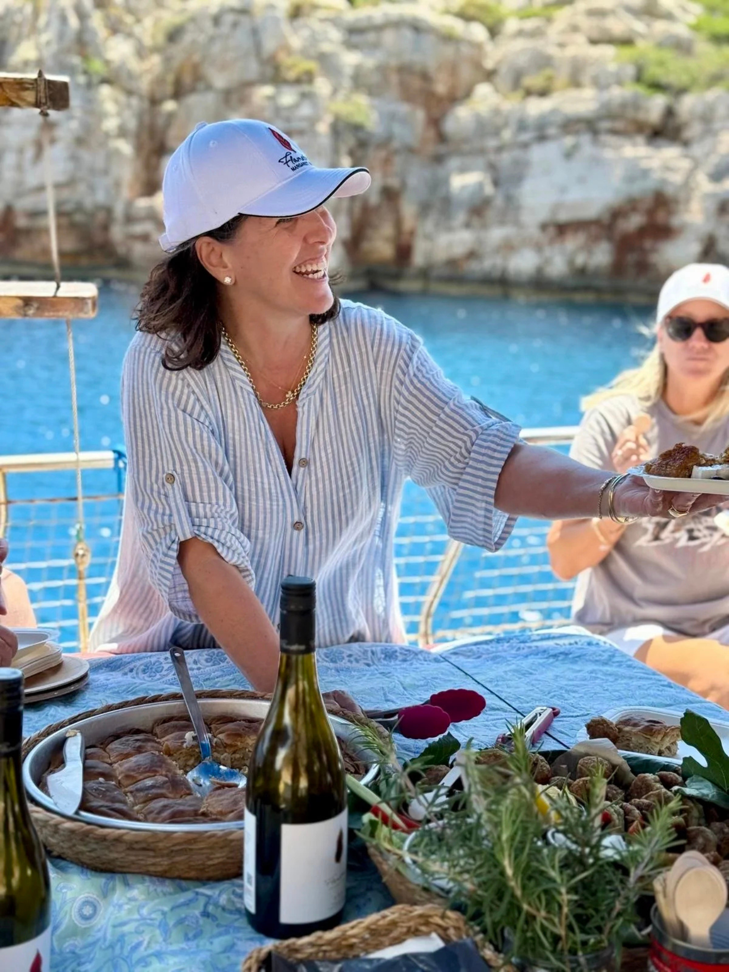 A woman wearing a striped shirt, white cap, and gold necklace is smiling and serving food at an outdoor gathering near a body of water with rocky cliffs in the background.